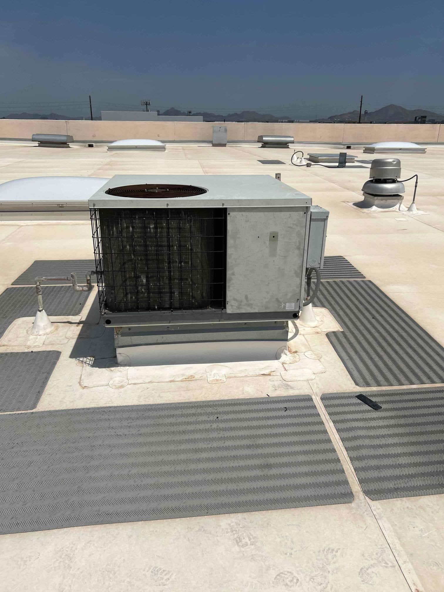 Rooftop air conditioning condenser unit on a flat roof, showing weathered condenser coil and protective grille under direct sunlight.