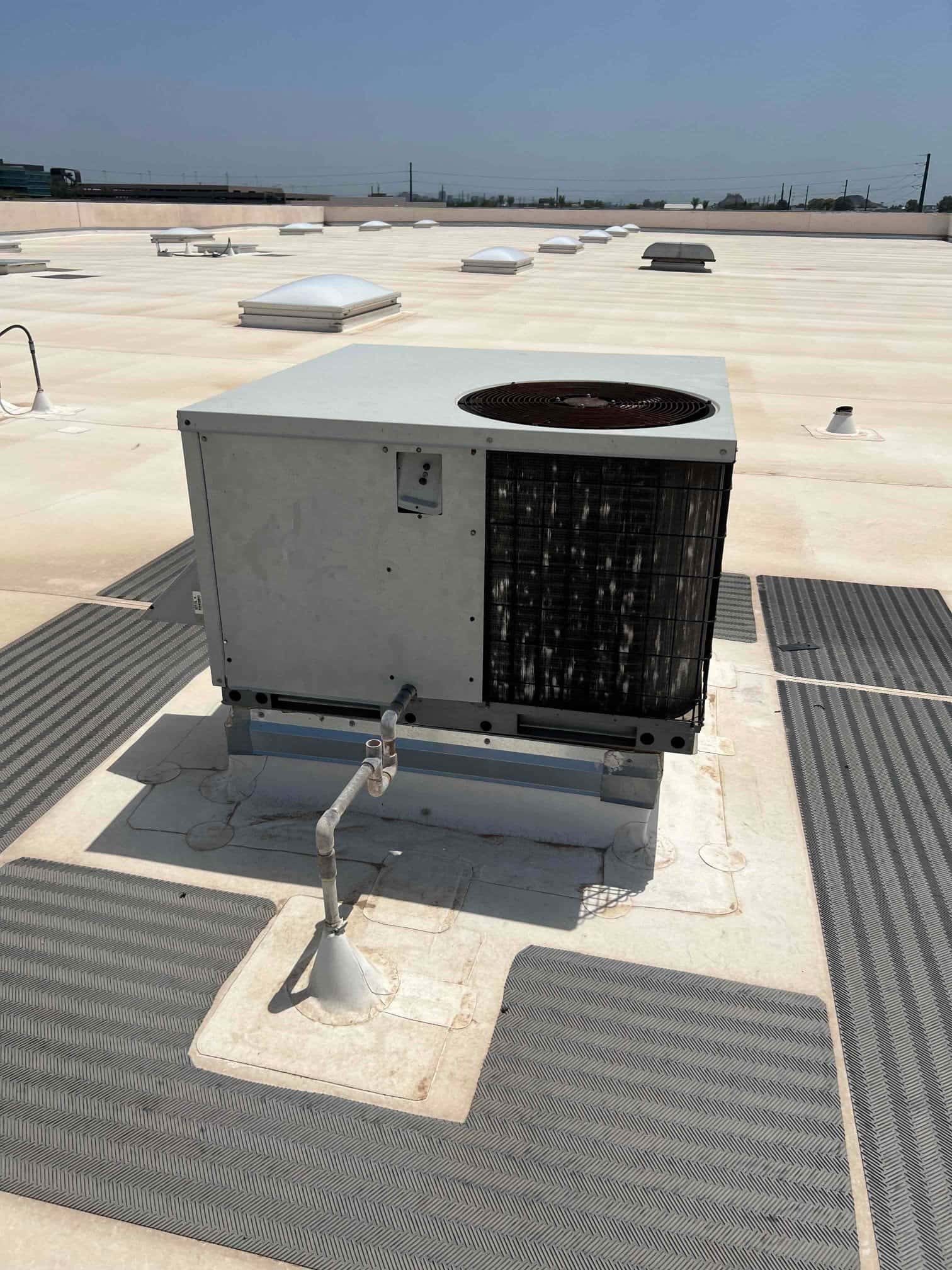 Rooftop air conditioning unit with exposed condenser coil and refrigerant piping on a flat commercial roof.