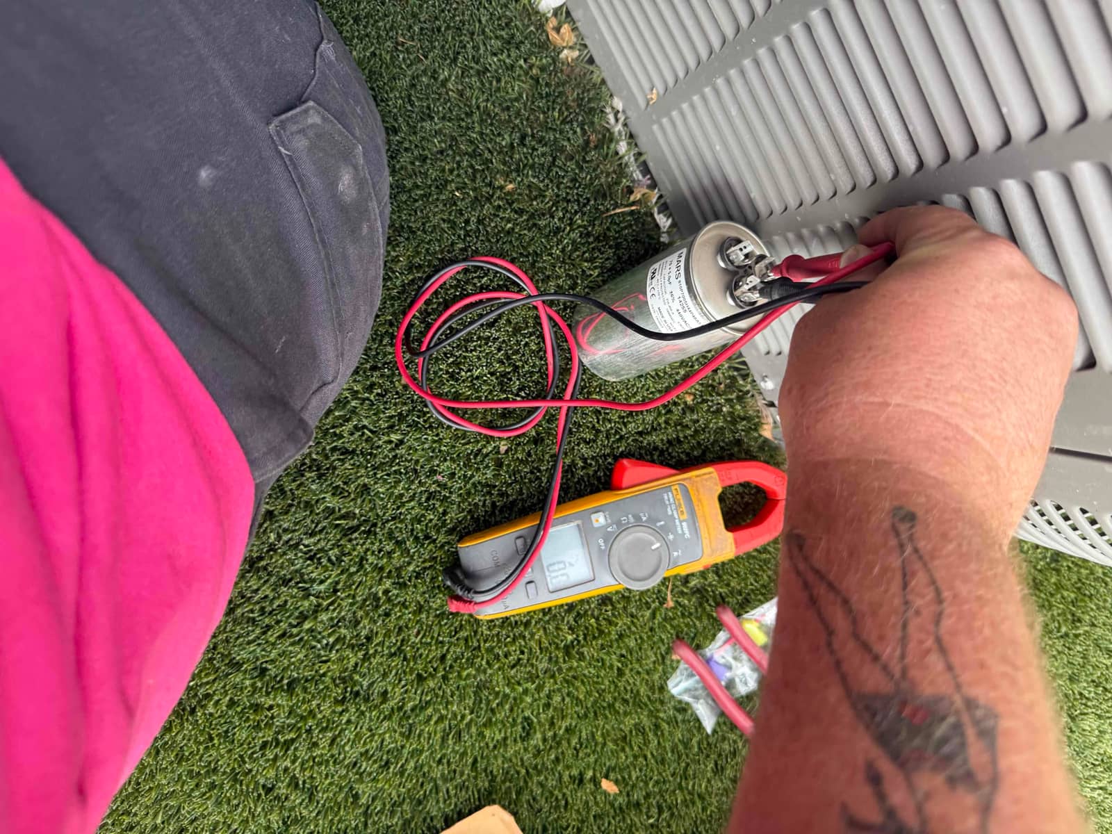 Technician measures a dual-run capacitor at the outdoor condenser using a clamp multimeter with test leads connected to the terminals.