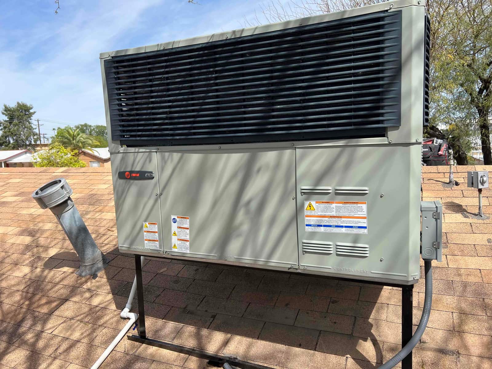 Rooftop packaged HVAC unit installed on stand with connected electrical whip and condensate drain, set on shingle roof.