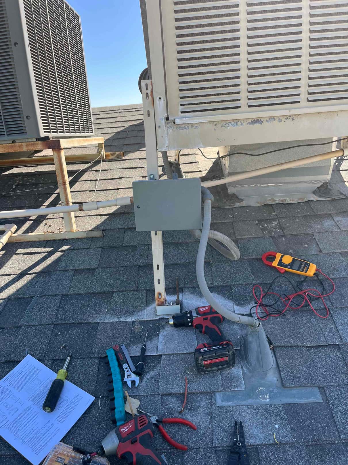 Digital multimeter rests on the roof near an open electrical junction box below a rooftop air conditioning unit.