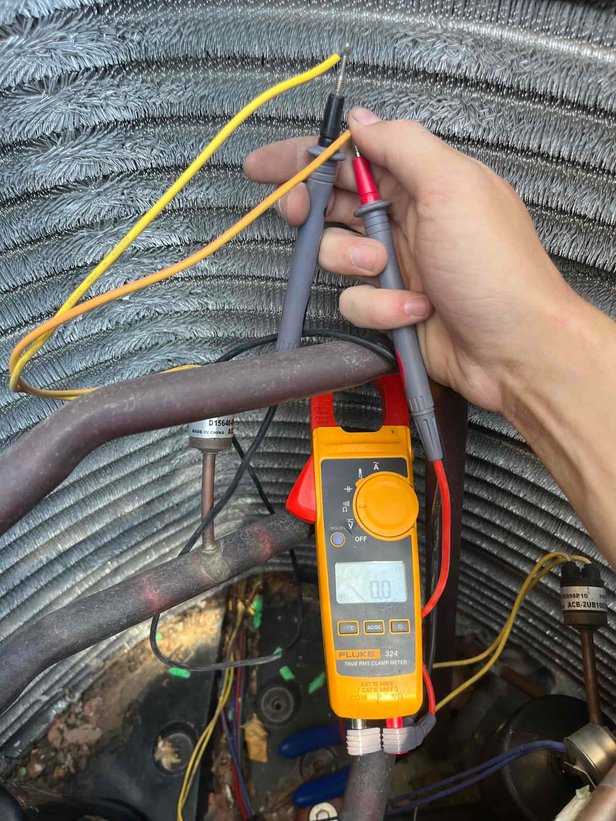 Digital multimeter set to volts shows 0.0 V with probes across low-voltage control wires inside the condenser.