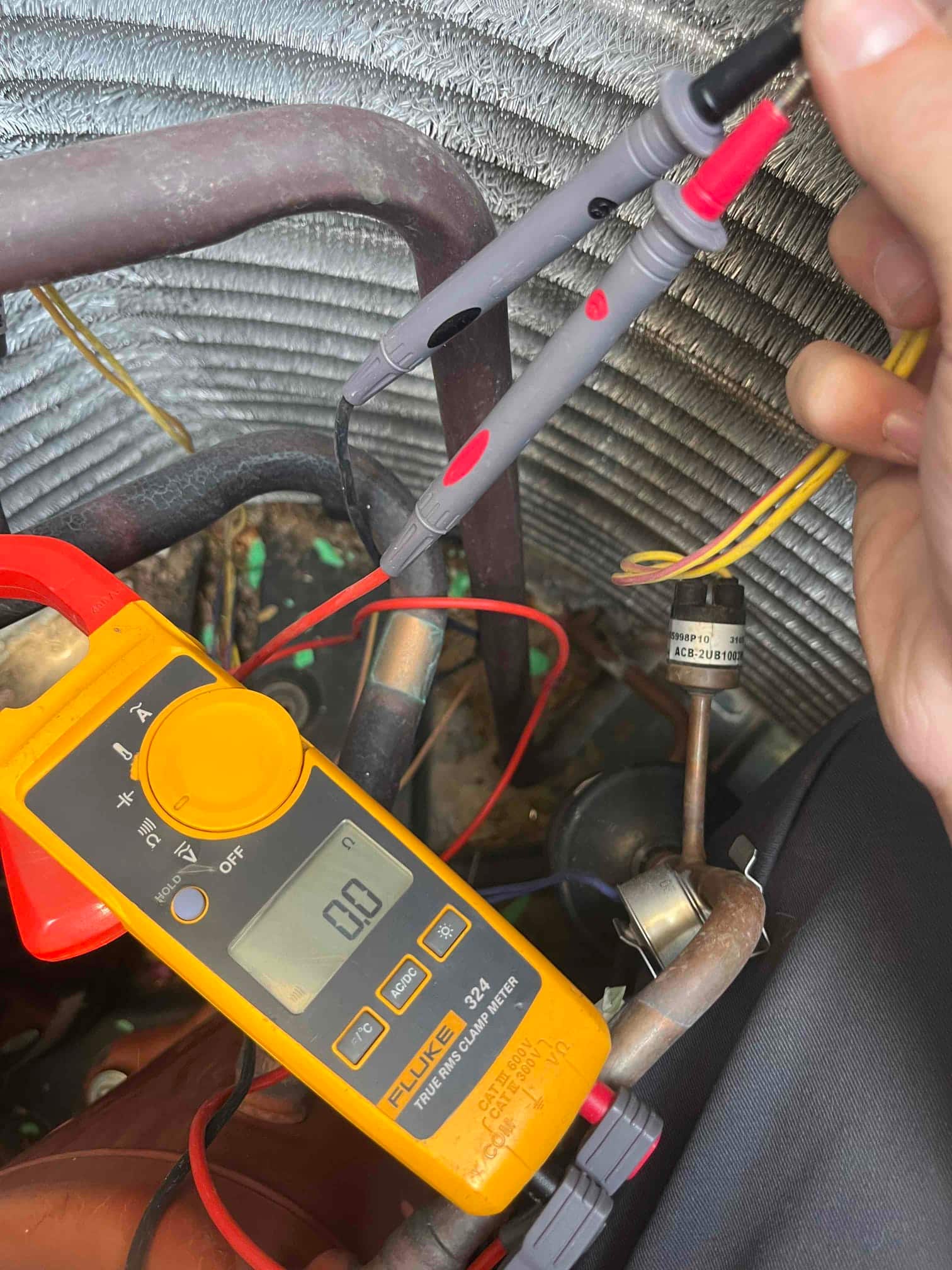 Digital multimeter shows 0.0 V with probes on yellow control wires beside pressure switch tubing and condenser coil.