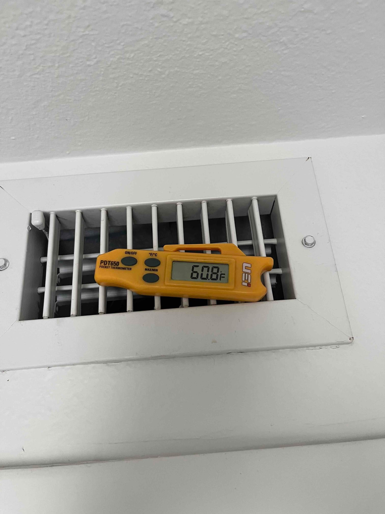Digital thermometer rests on a supply vent grille, displaying supply air temperature of 60.8 °F.
