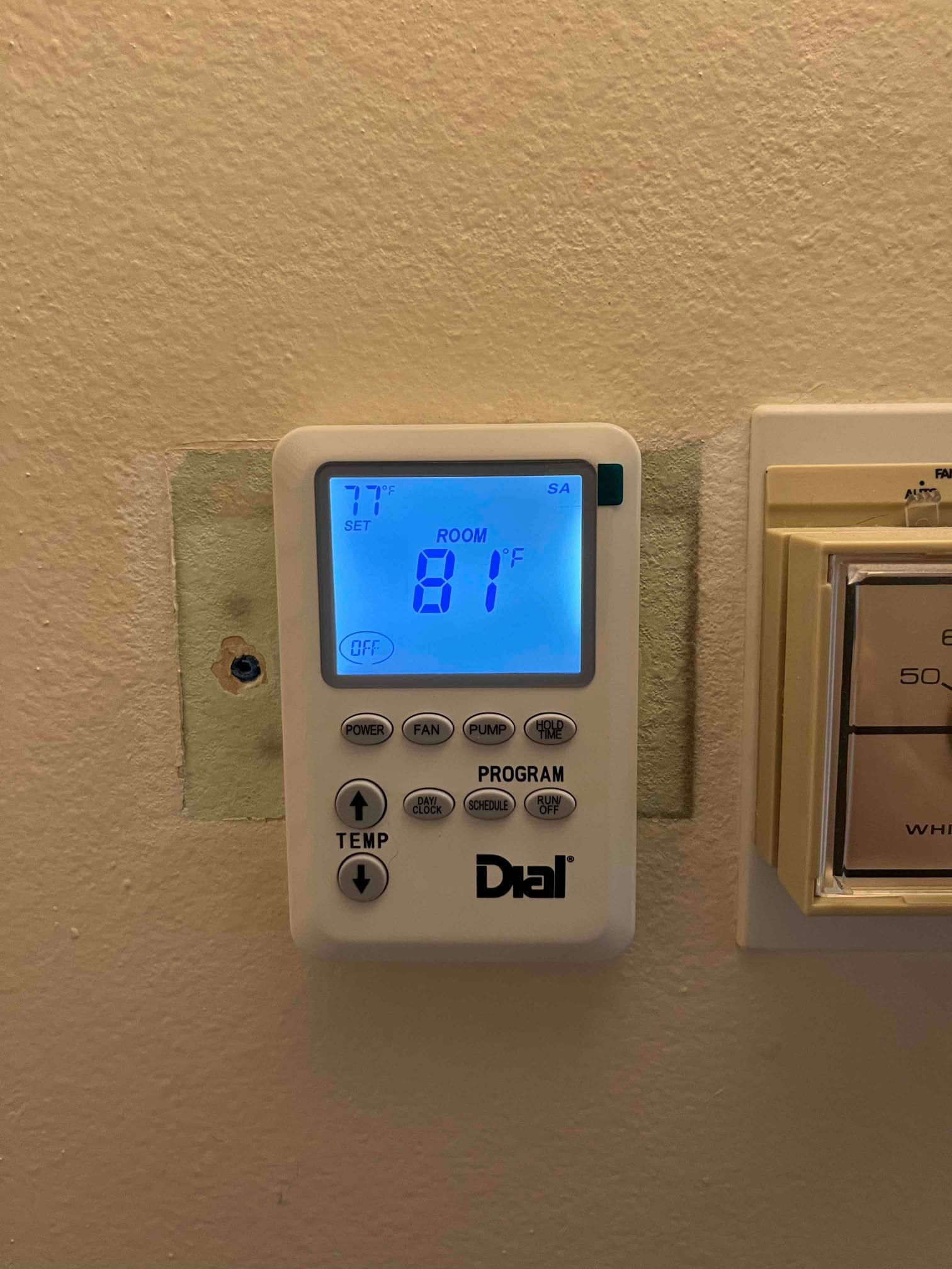 Digital thermostat on wall displays room temperature 81°F and setpoint 77°F, with backlight on and adjacent control plate.
