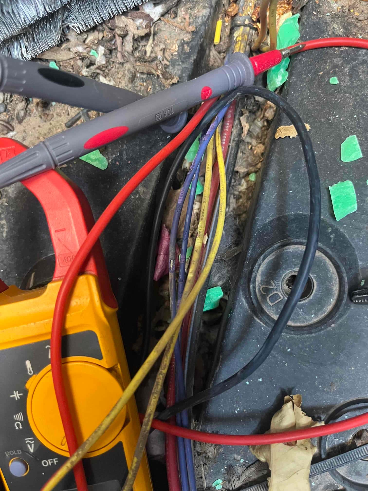 Handheld multimeter with clamp and test leads positioned on HVAC wiring harness near the outdoor unit base.
