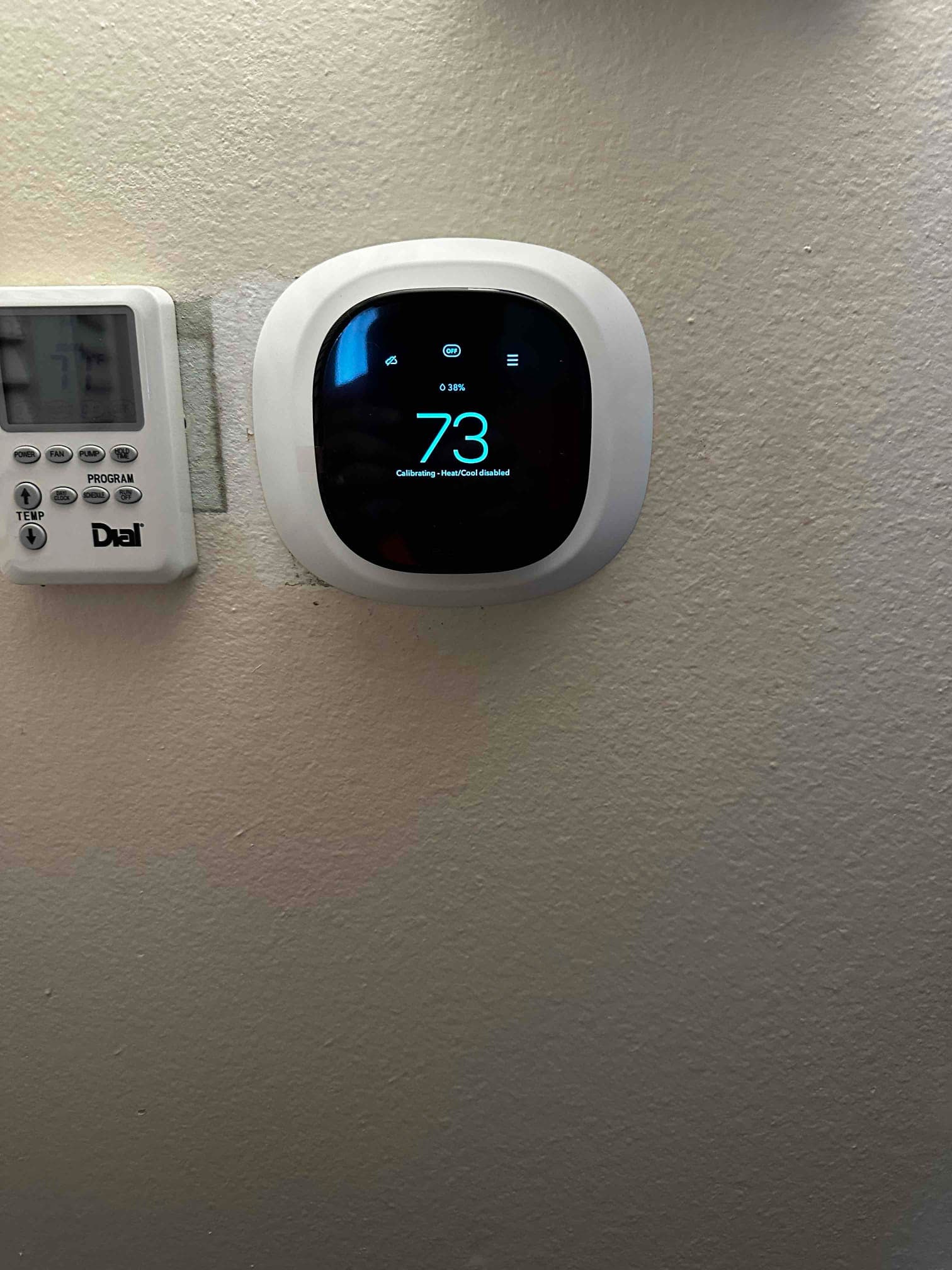 New smart thermostat mounted and powering up, displaying 73 degrees alongside the existing programmable thermostat on the wall.