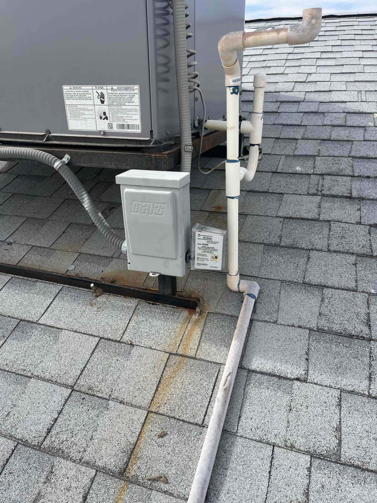 Outdoor HVAC unit on a roof with electrical disconnect, condensate piping, and rust staining beneath the equipment.