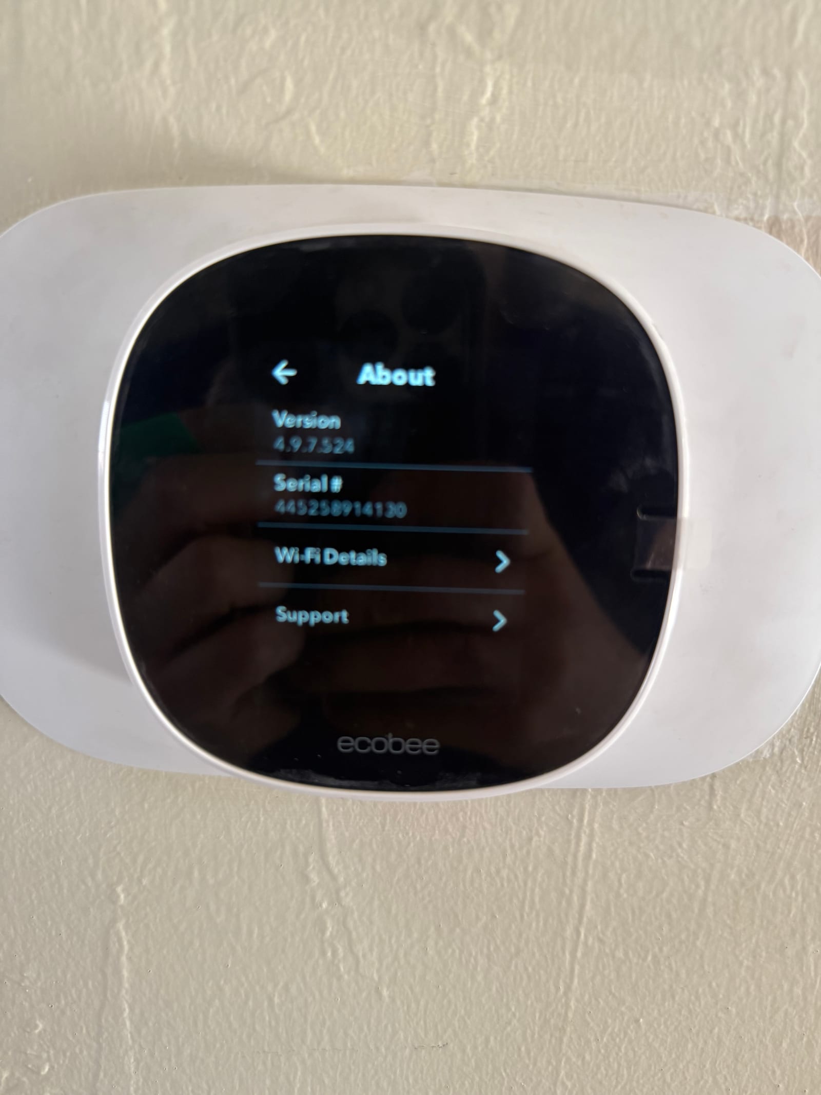 Smart thermostat mounted on wall displaying the About menu with version, serial, Wi‑Fi details, and support options.