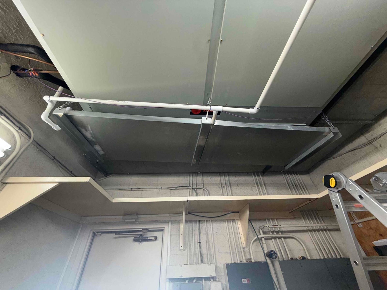 Suspended indoor air handler installed with new PVC condensate drain piping and supporting hangers in a mechanical room.