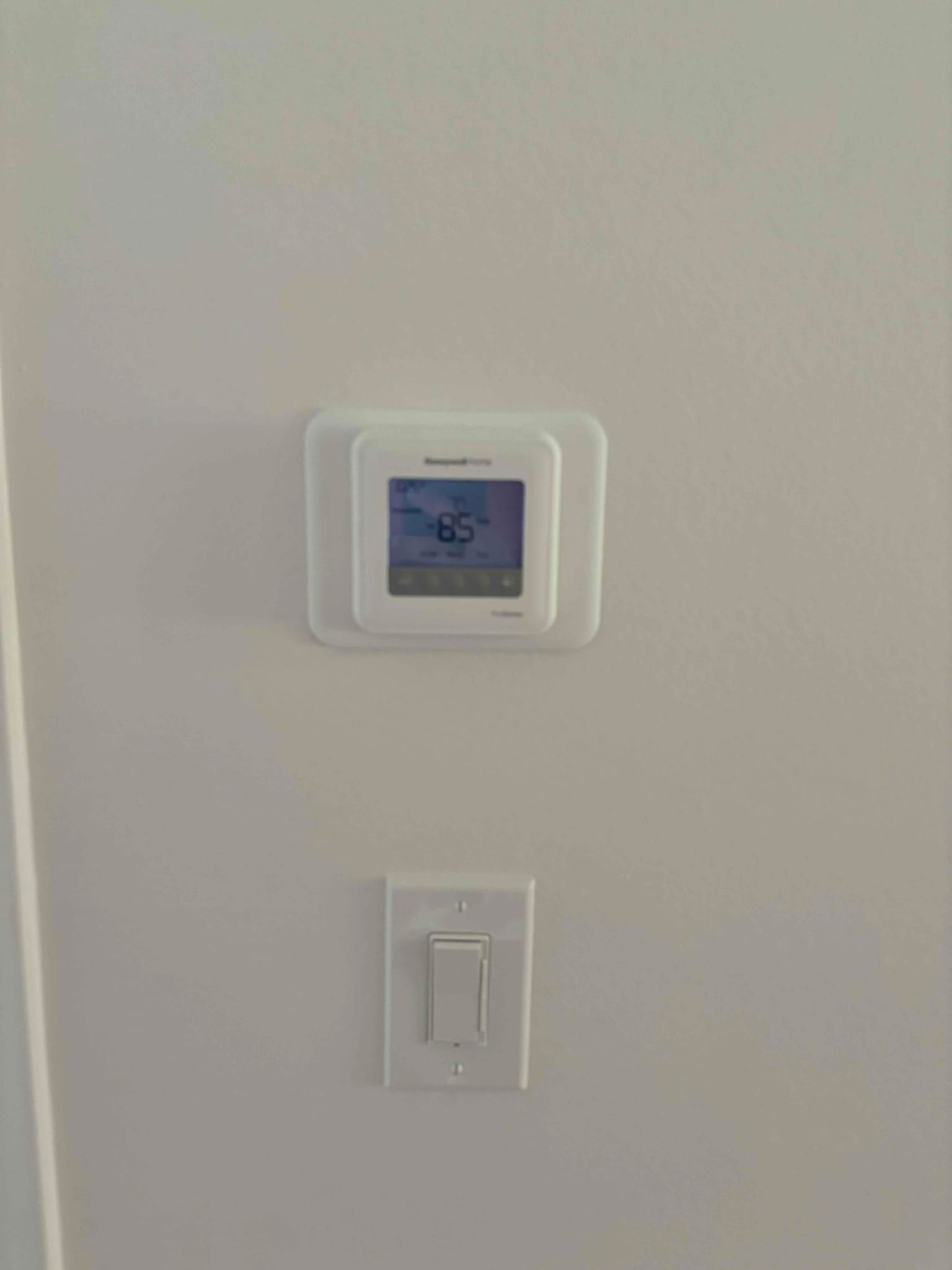 Wall-mounted digital thermostat installed above a light switch, screen illuminated and displaying a temperature reading.