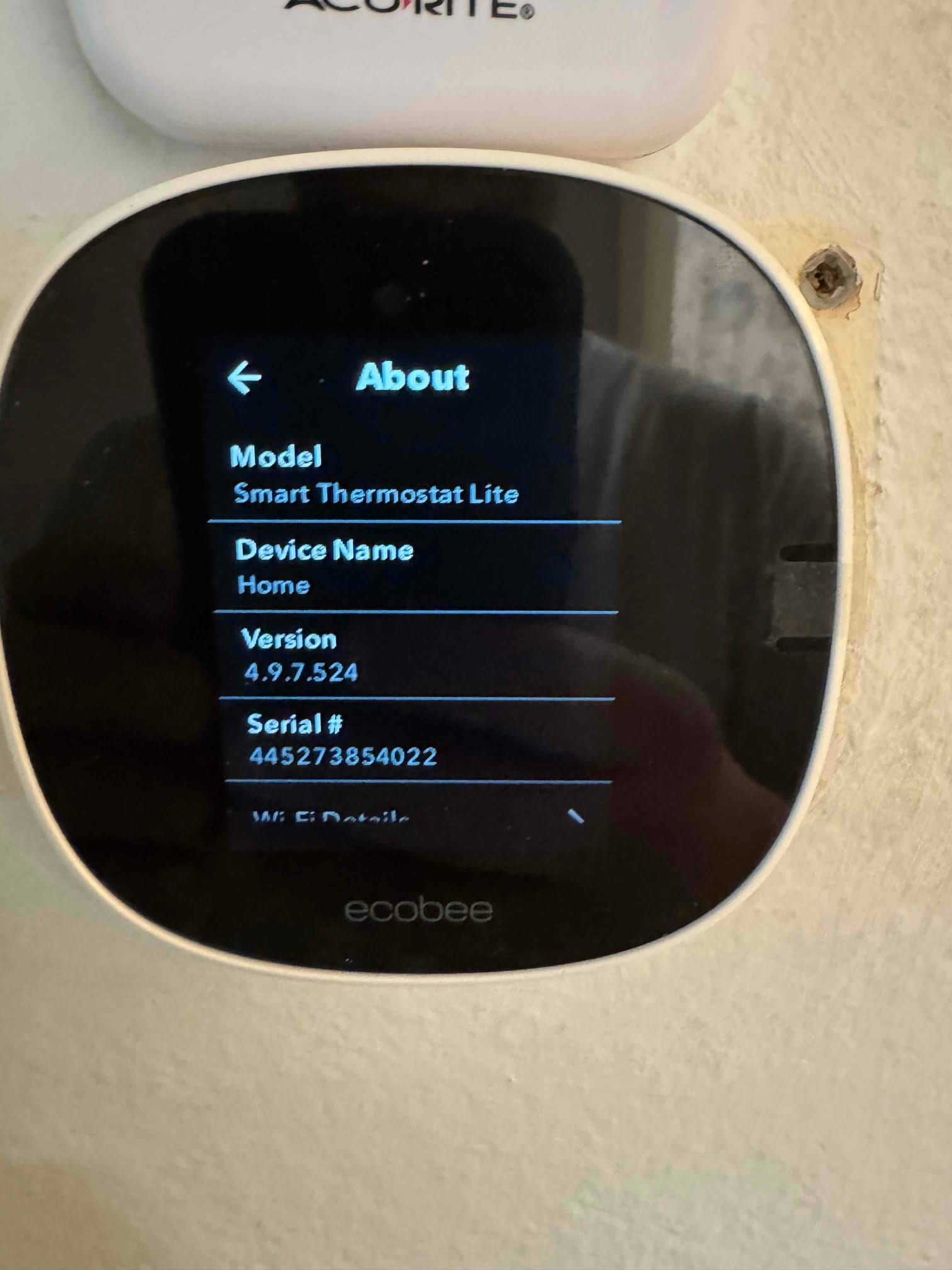 Wall-mounted smart thermostat displays About menu during setup, confirming model, device name, version, and connectivity details.