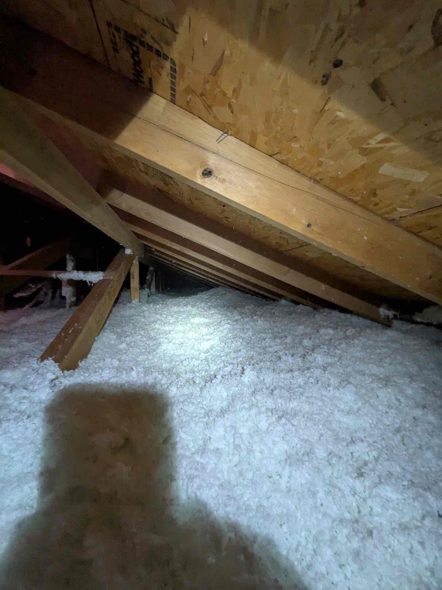 Attic space with blown-in insulation under roof rafters, illuminated by a work light during HVAC installation access.