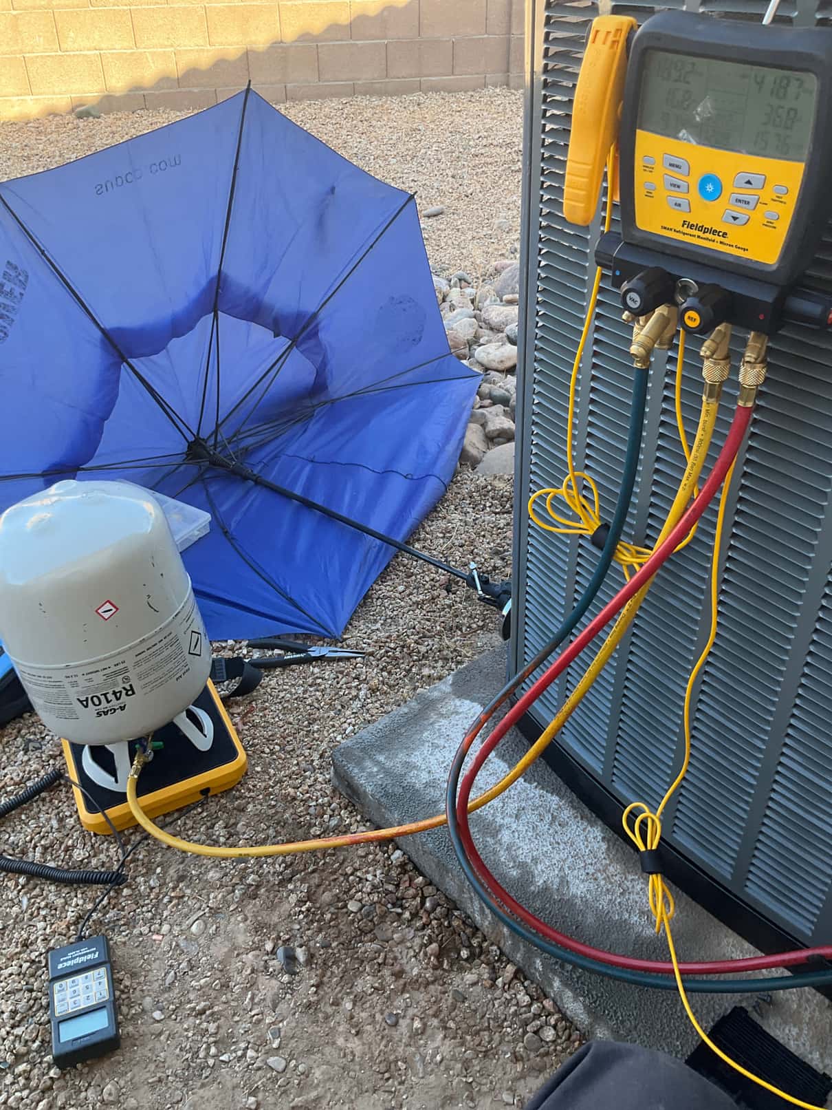 Digital manifold gauges on the outdoor unit display different high and low pressures while charging from a refrigerant cylinder on a scale.