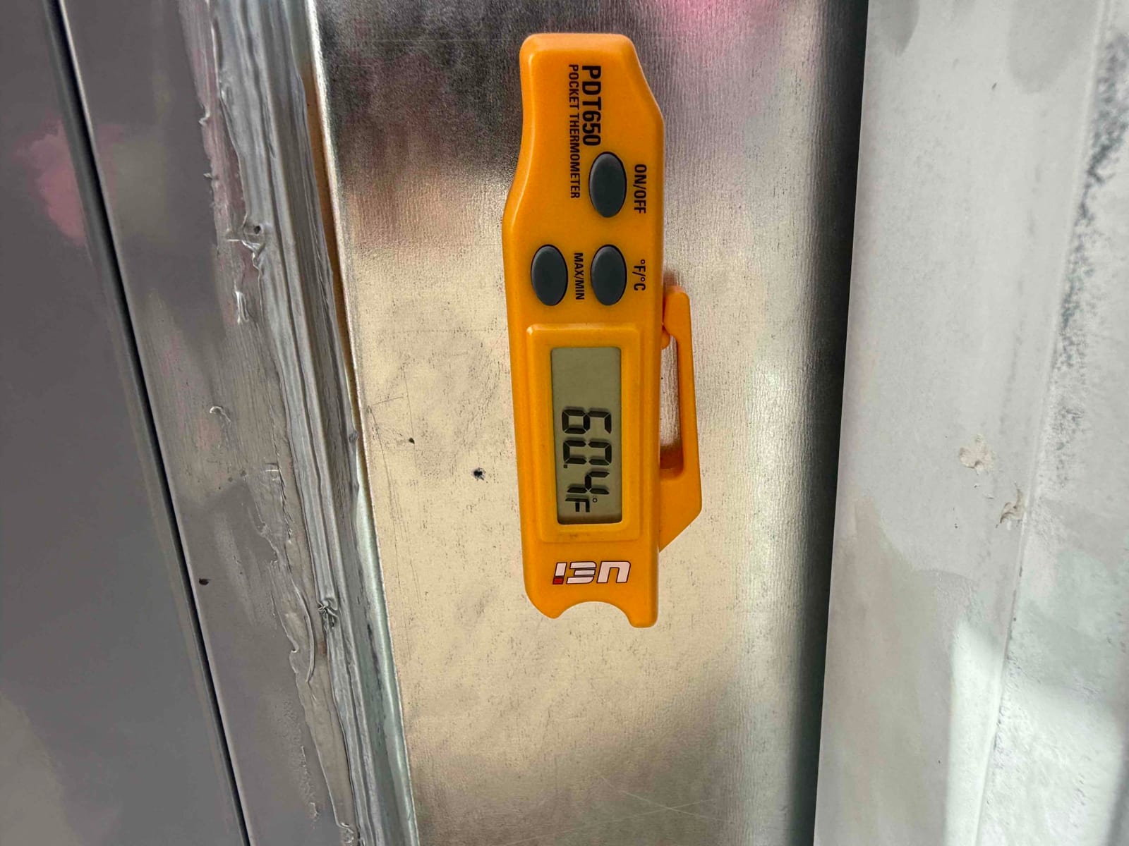 Digital pocket thermometer shows 60.4°F while measuring air temperature at the metal duct surface during commissioning.