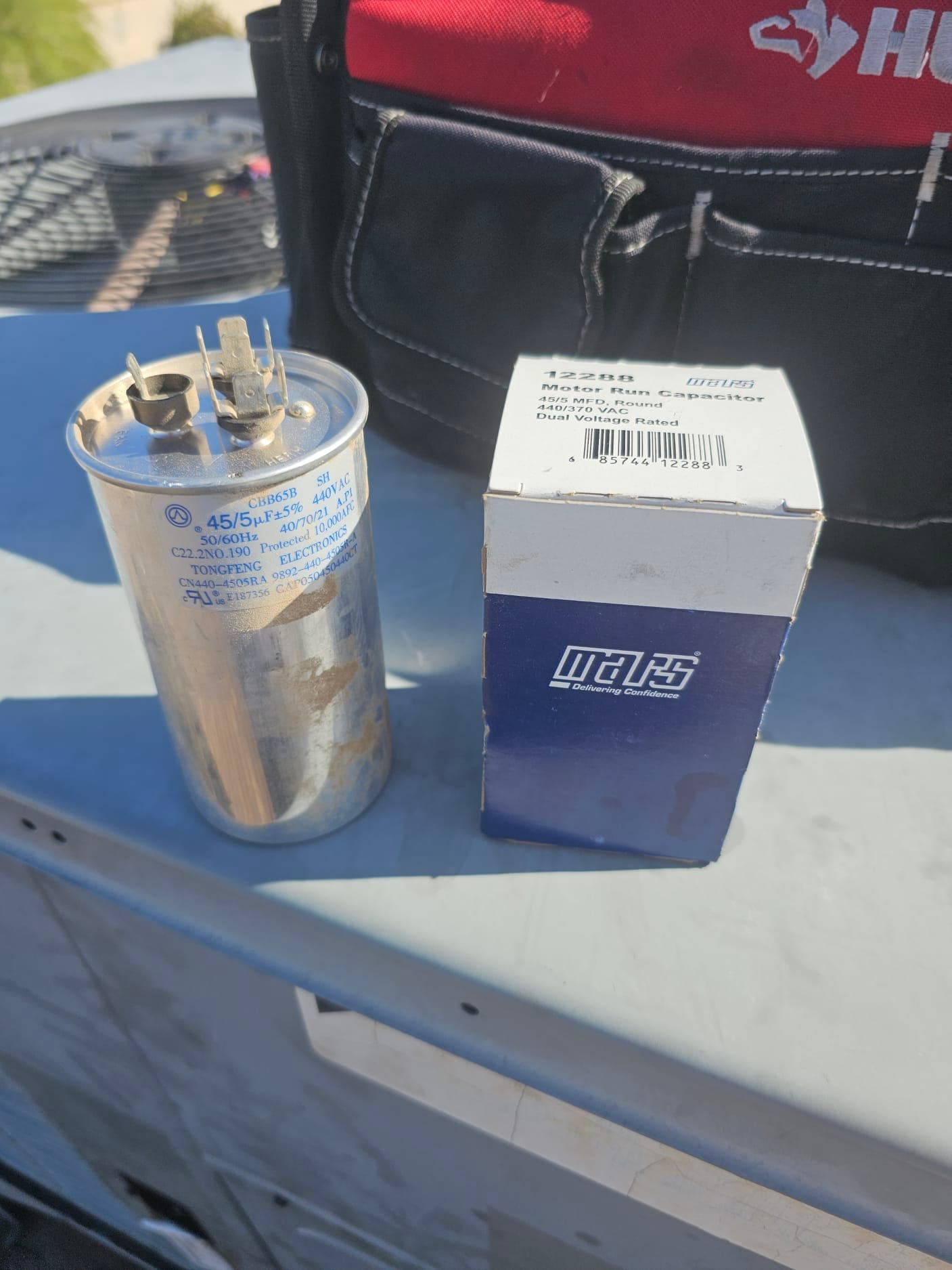 Dual-run capacitor labeled 45/5 μF with wiring terminals exposed, placed beside a boxed replacement on the condenser.