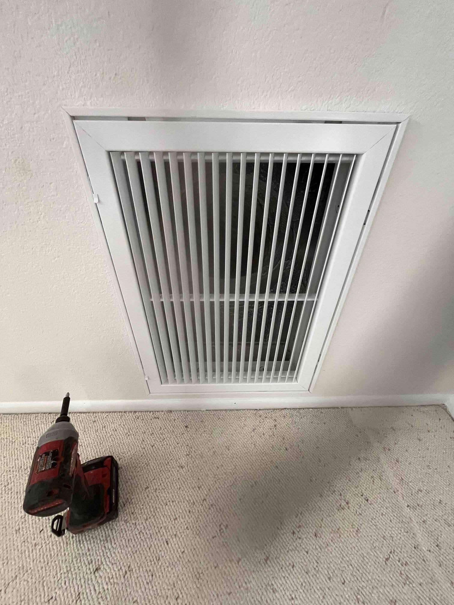 New return air grille installed on interior wall, with power drill resting on carpet after filter frame replacement.