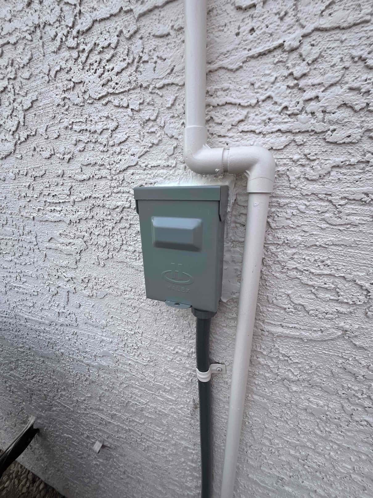 Weatherproof HVAC disconnect and sealed conduit installed on exterior wall beside painted PVC condensate drain.