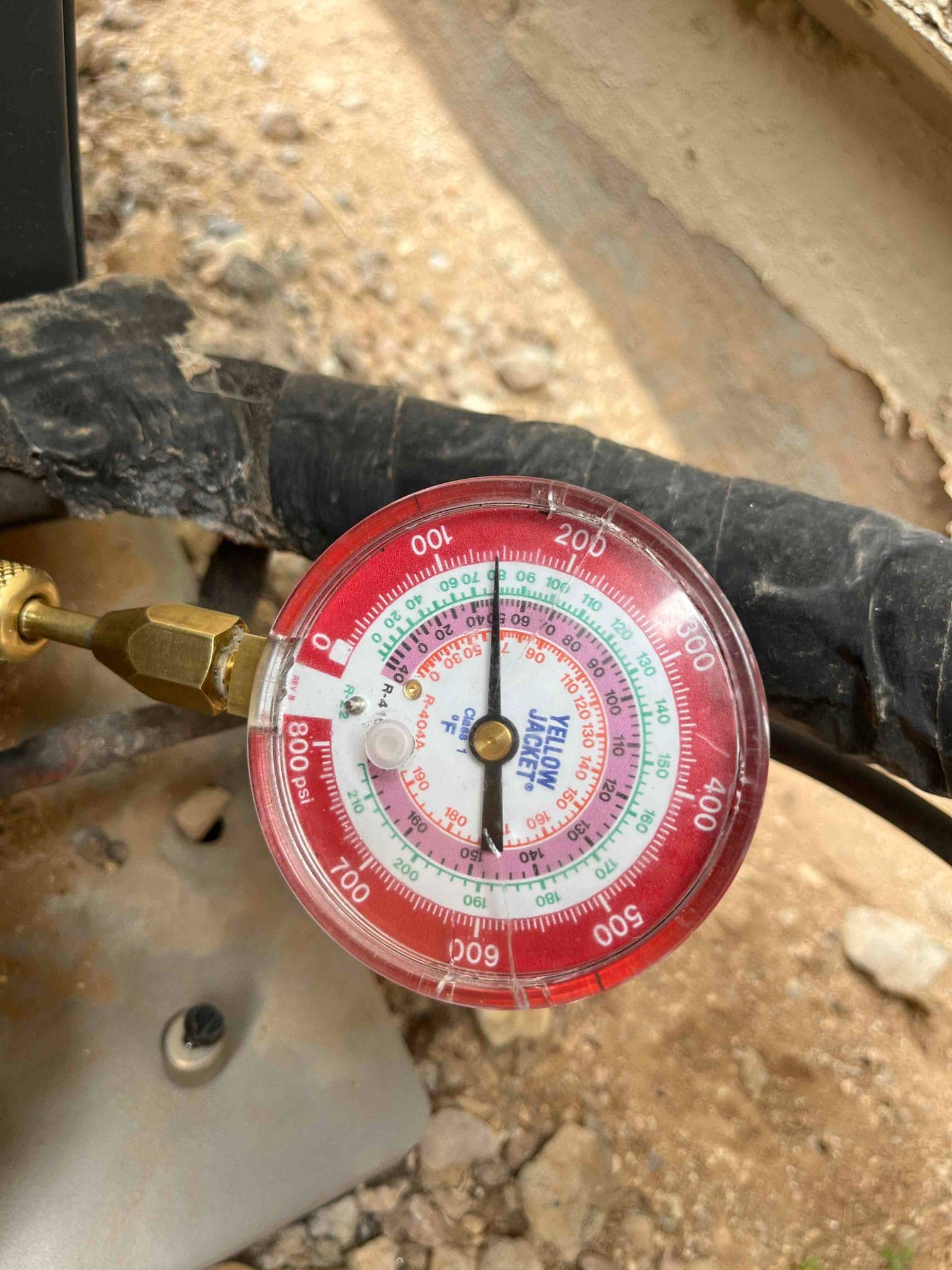 Analog refrigerant gauge connected to a service port on outdoor HVAC equipment, showing pressure scales with needle near the low-pressure range.