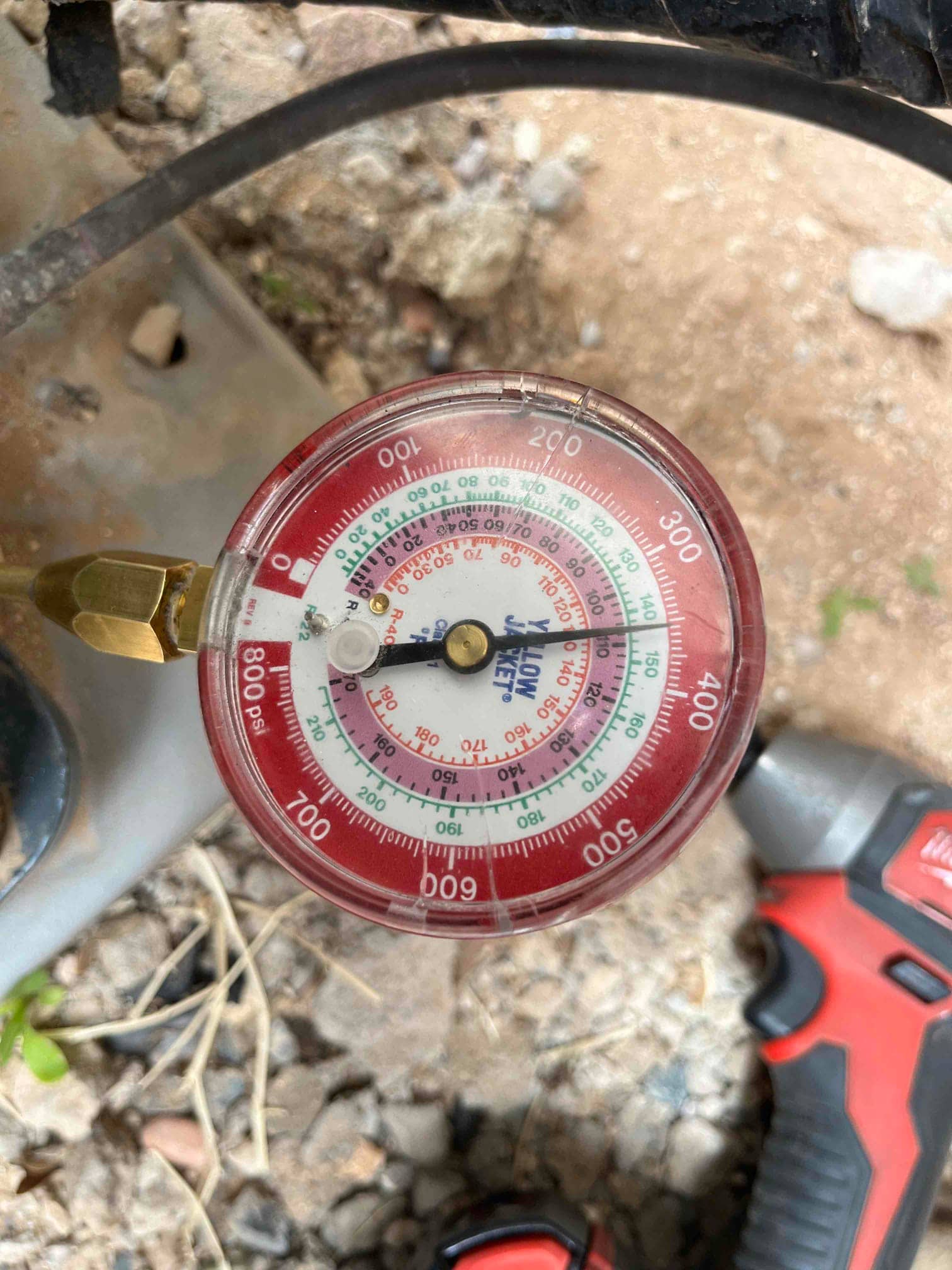 Analog refrigerant manifold gauge in use on an outdoor unit, showing multiple pressure scales and temperature conversion rings.
