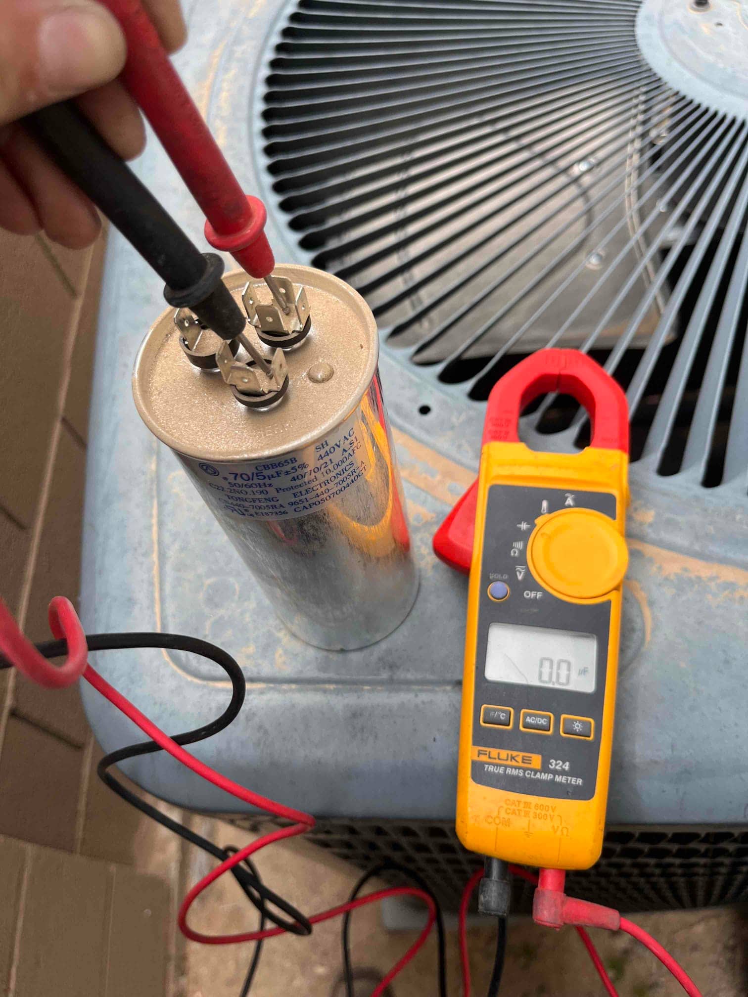 Clamp meter display shows 0.0 V while test leads contact the dual-run capacitor terminals near the outdoor condenser.