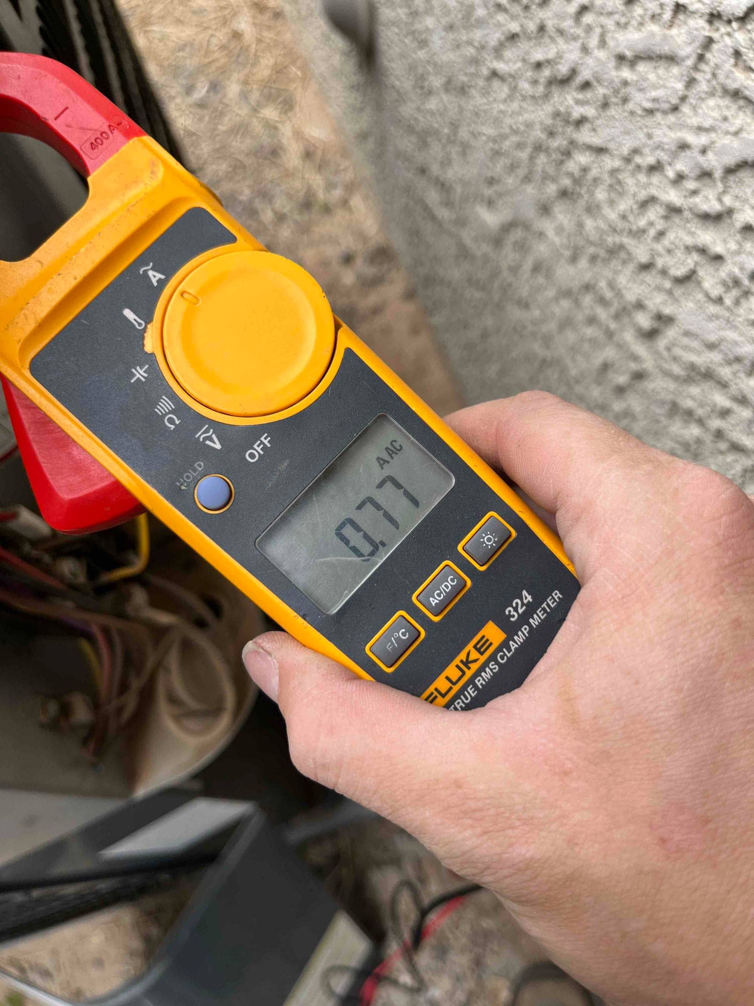 Clamp meter displays 0.77 A AC with the jaw clamped on a single conductor near the outdoor unit.