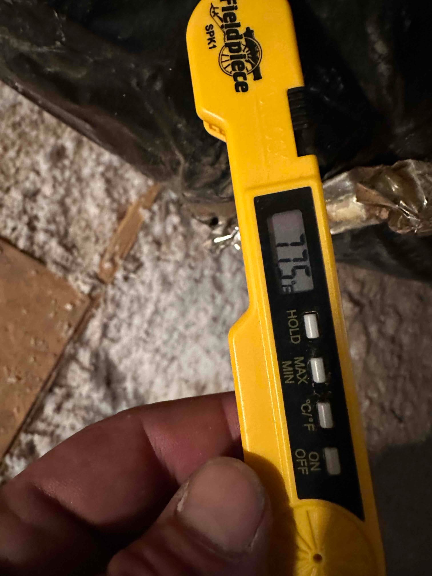 Digital thermometer probe reads 77.6°F while measuring air temperature at the duct during commissioning checks.