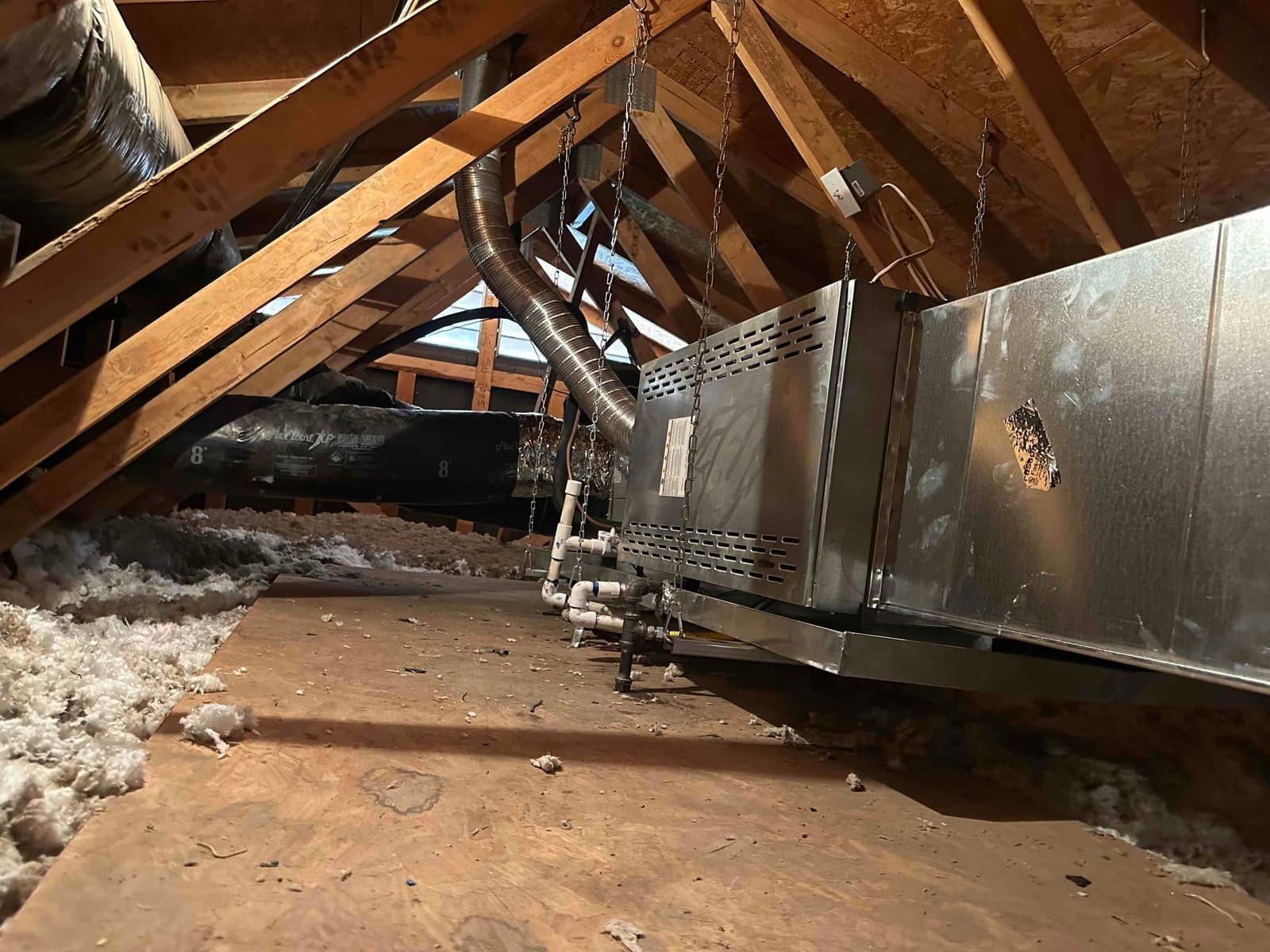 Horizontal attic air handler hung from rafters with insulated duct connections and PVC condensate drain piping above a secondary drain pan.