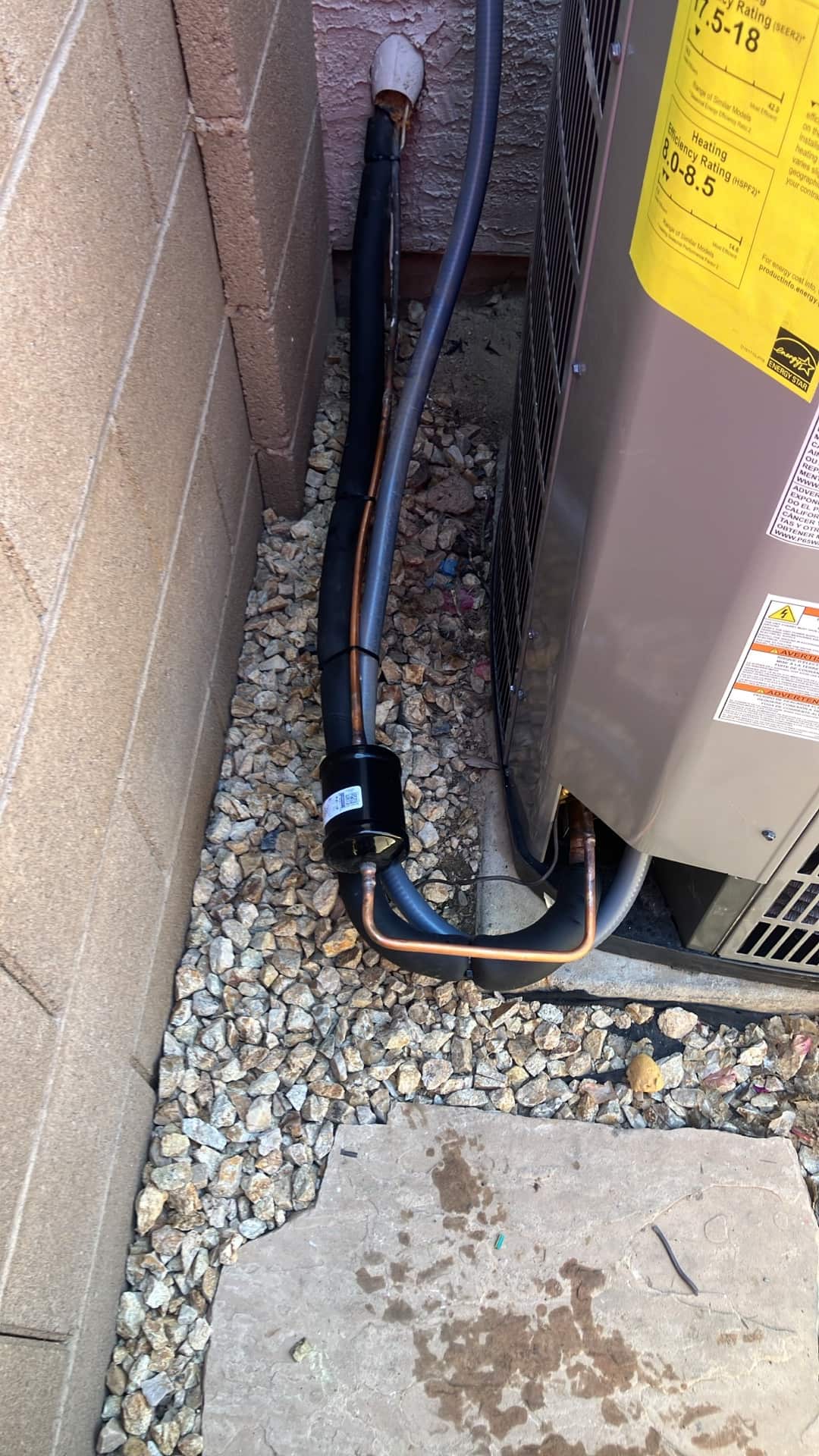New outdoor condenser connections with insulated suction line, liquid line filter-drier, and sealed wall penetration routed over decorative rock.