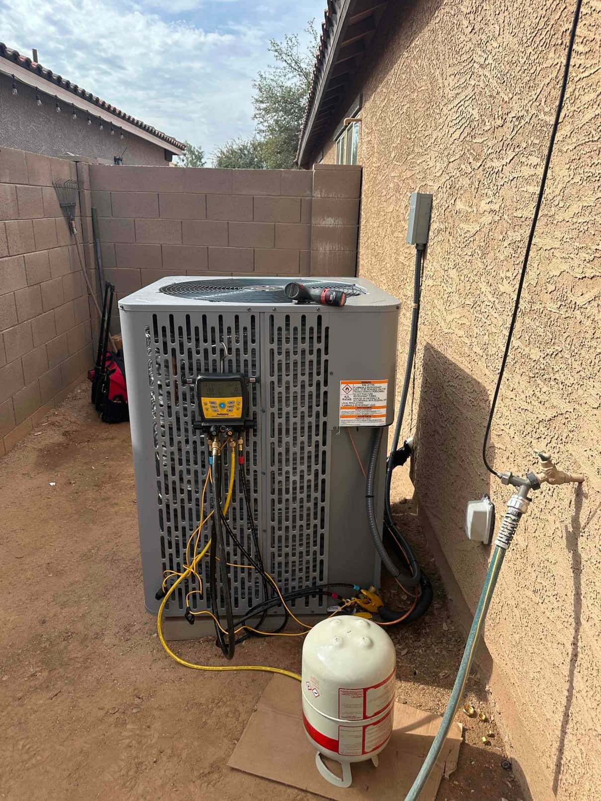 New outdoor heat pump condenser set on a pad with manifold gauge connected and refrigerant cylinder staged beside the unit.