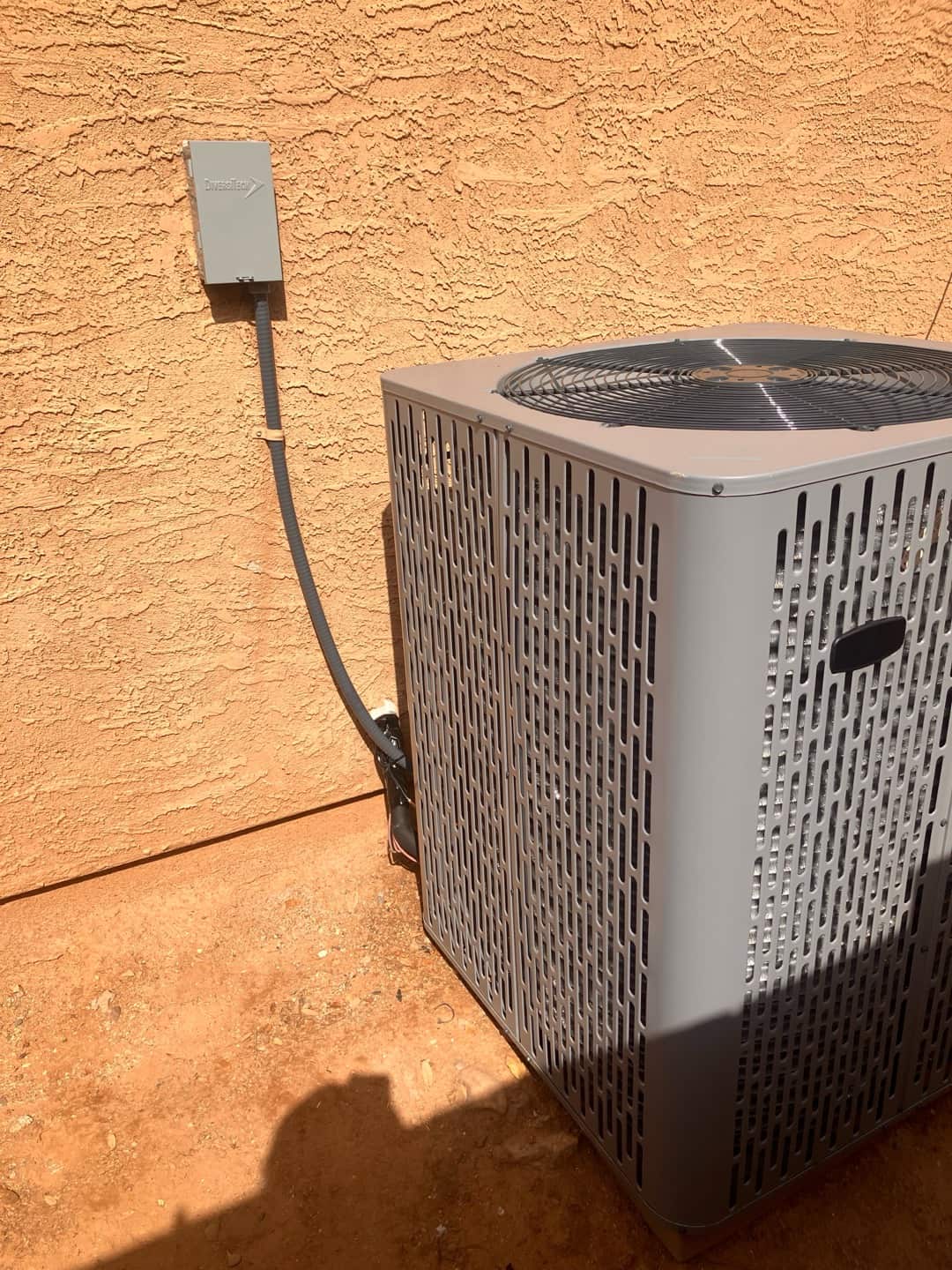 Outdoor heat pump condenser set beside stucco wall with weatherproof disconnect and flexible electrical whip neatly secured.