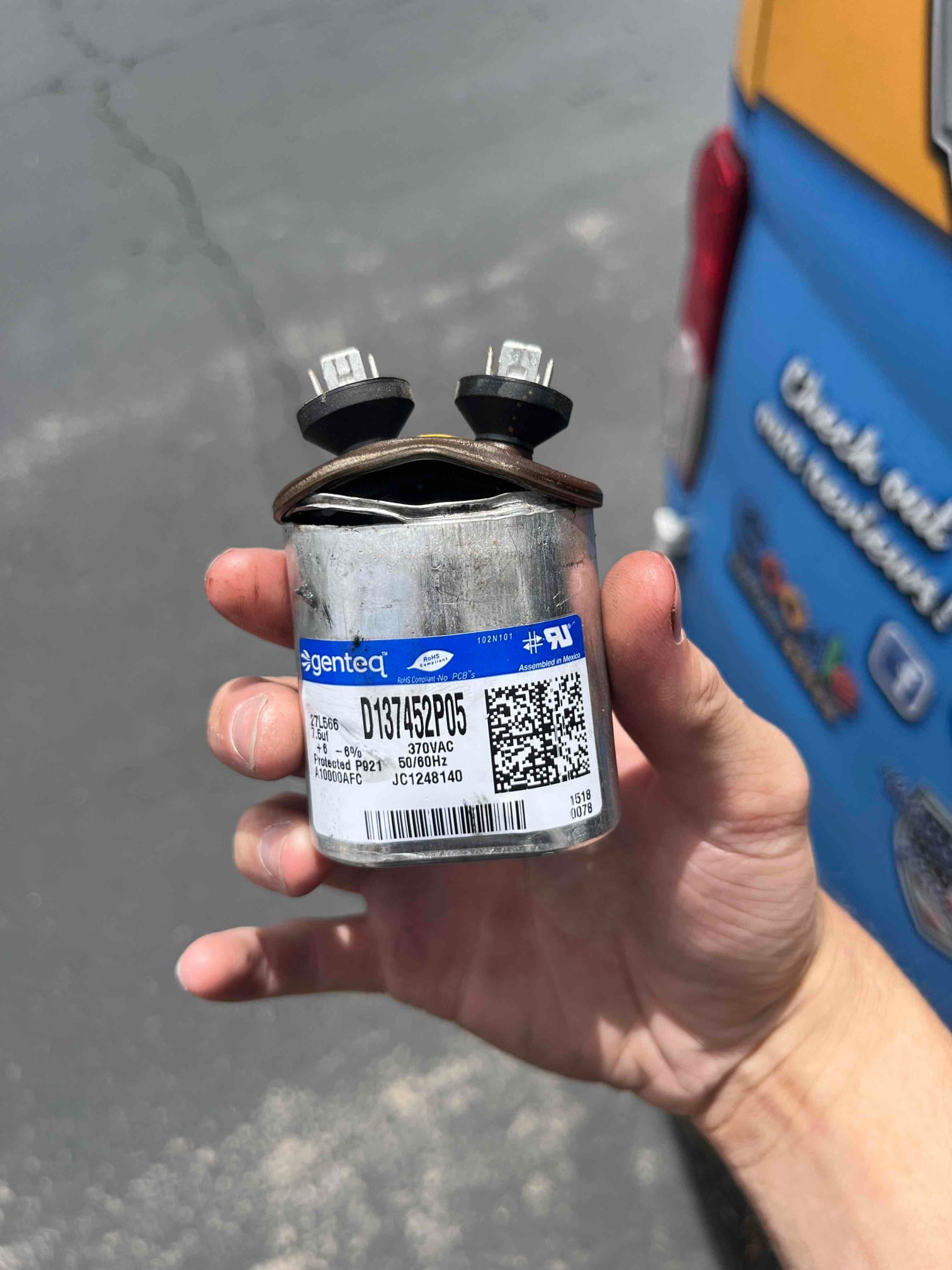 Removed run capacitor with swollen, bulged top and deformed case, showing failure. Technician holds the damaged component outdoors.