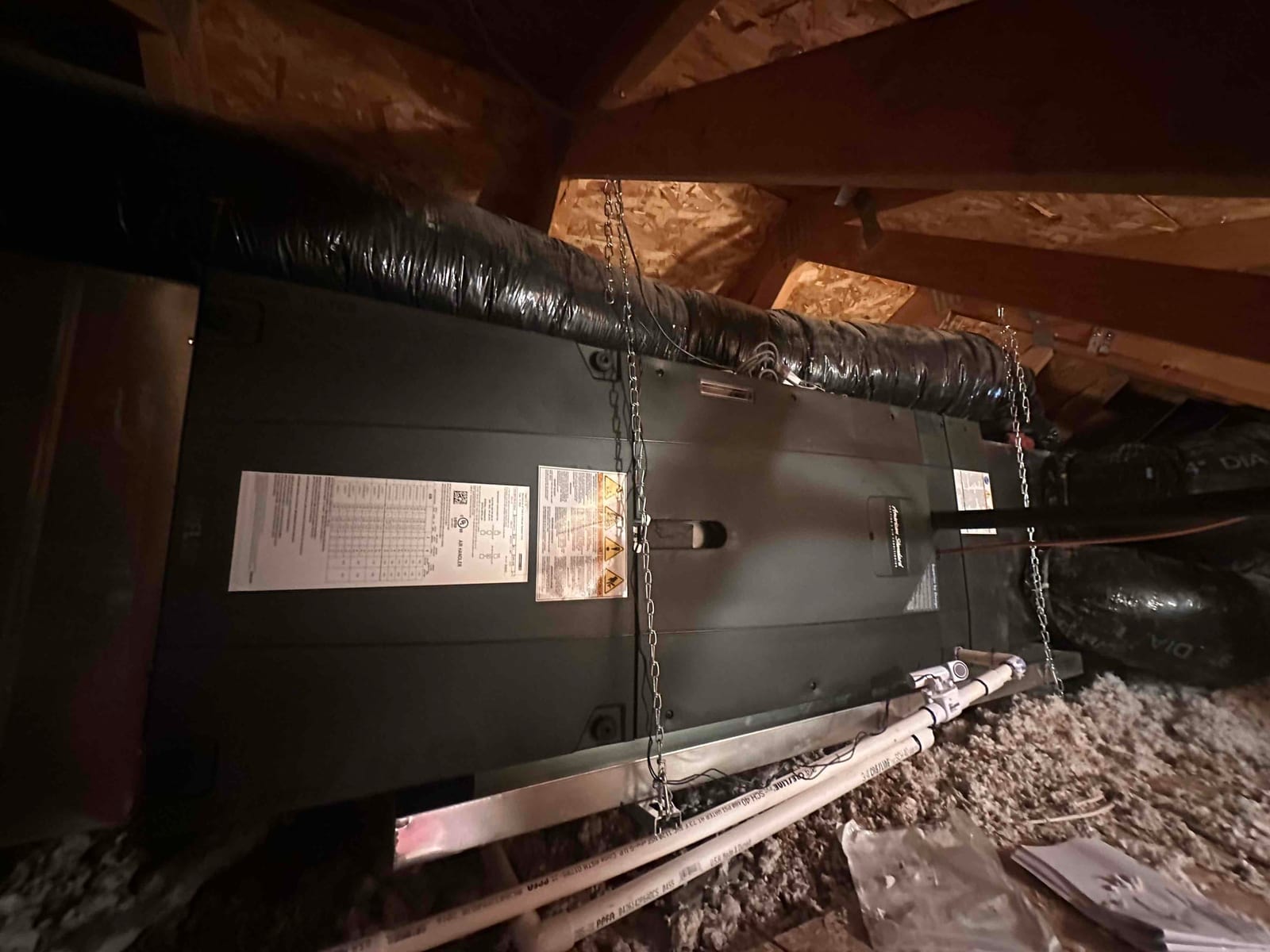 Suspended air handler installed in attic with insulated duct connections, condensate drain piping, and hanging chains secured to rafters.