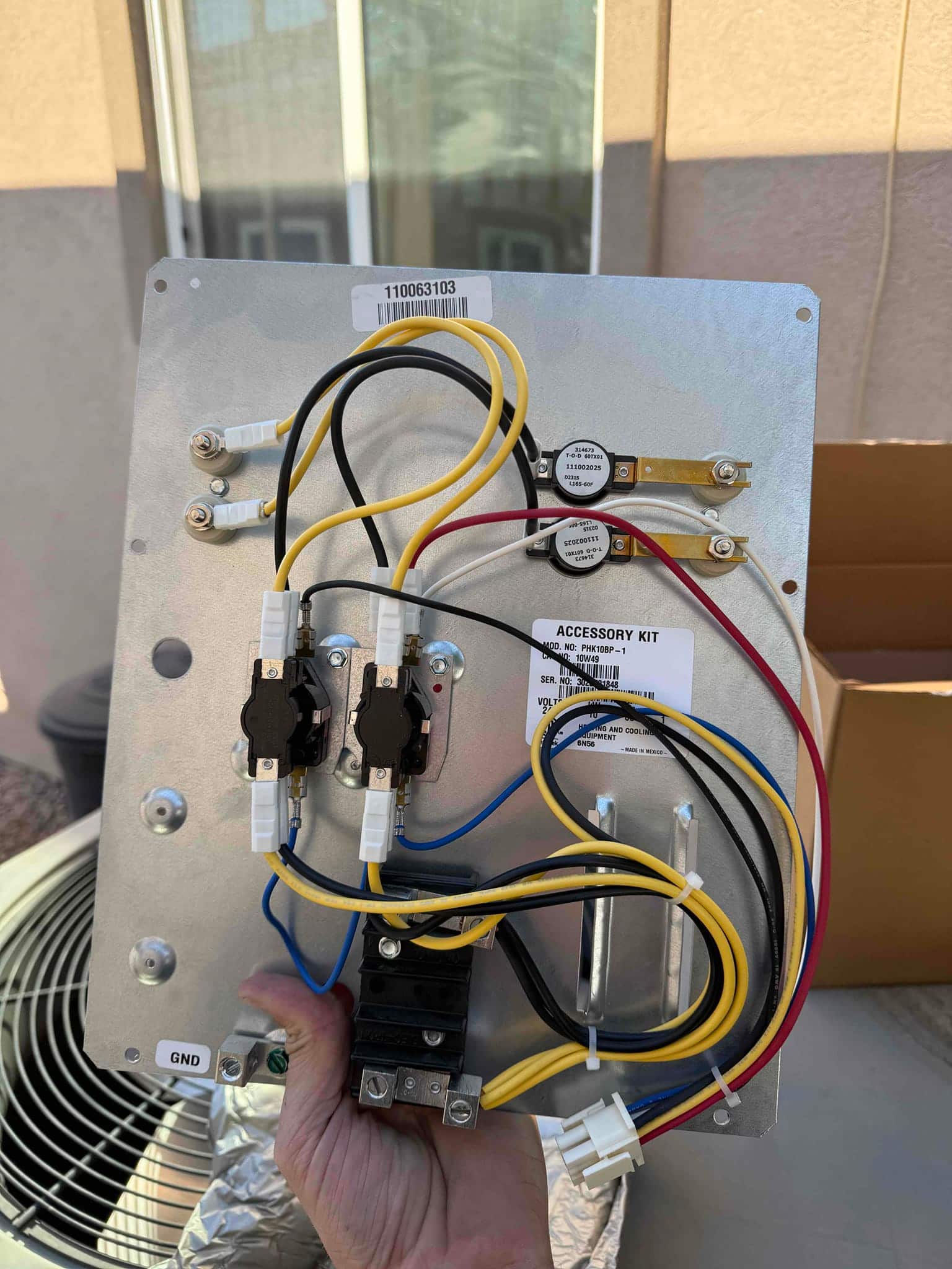 Technician holds an electric heat strip assembly with wired relays, limit switches, and terminal connections ready for installation.