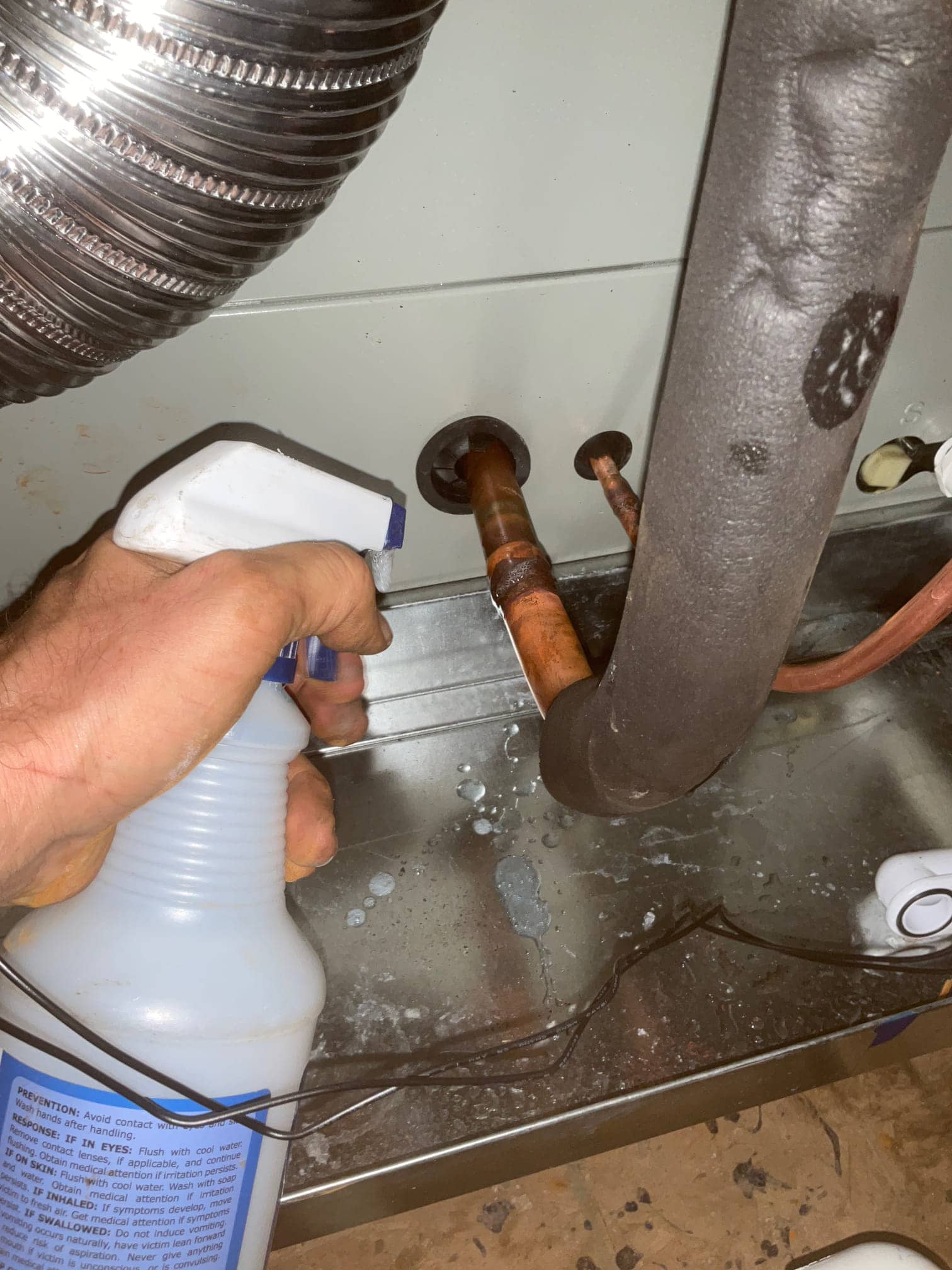 Technician sprays leak-detection solution on copper refrigerant lines at the indoor unit penetration during installation.