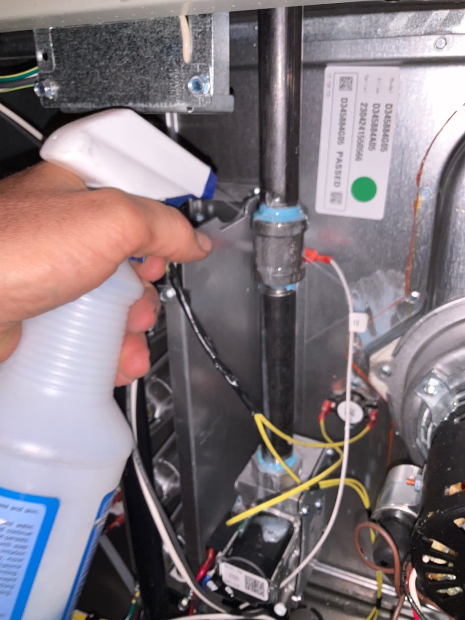 Technician sprays leak-detection solution on gas piping and fittings inside a furnace compartment with wiring and components visible.