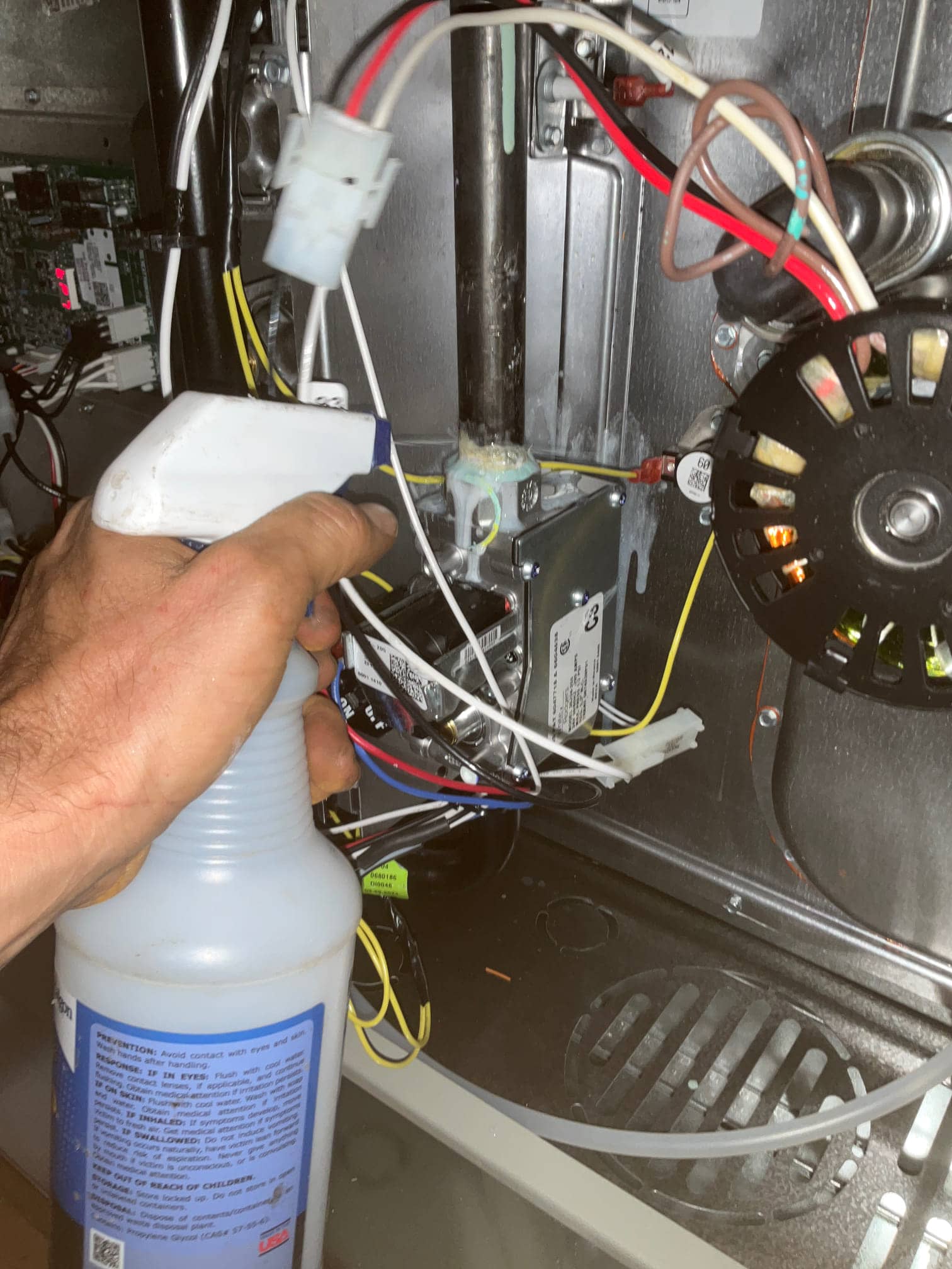 Technician sprays leak-detection solution inside a furnace cabinet near wiring, gas valve area, and inducer motor assembly.