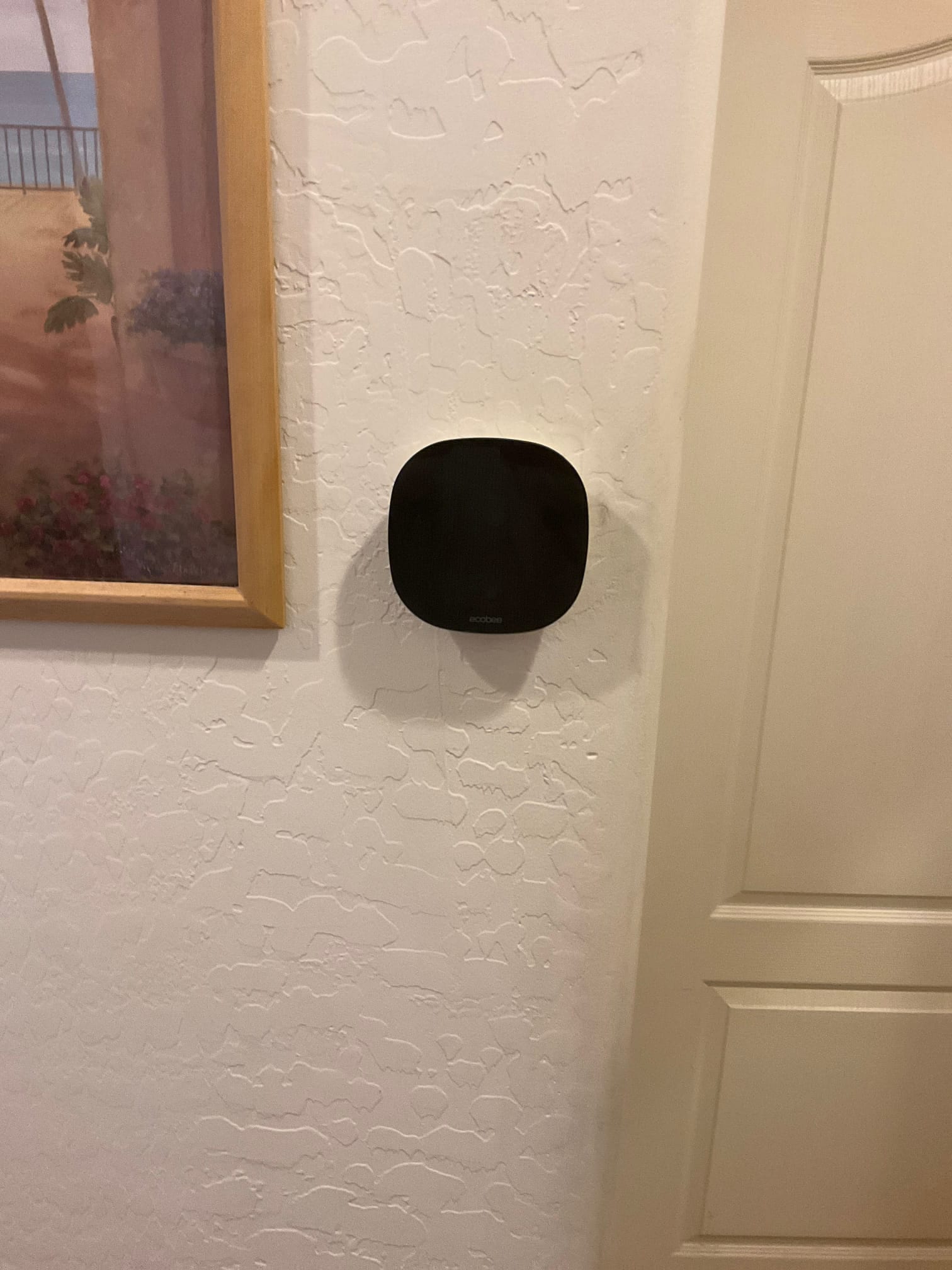 Wall-mounted smart thermostat installed on a finished interior wall adjacent to a door and framed artwork.