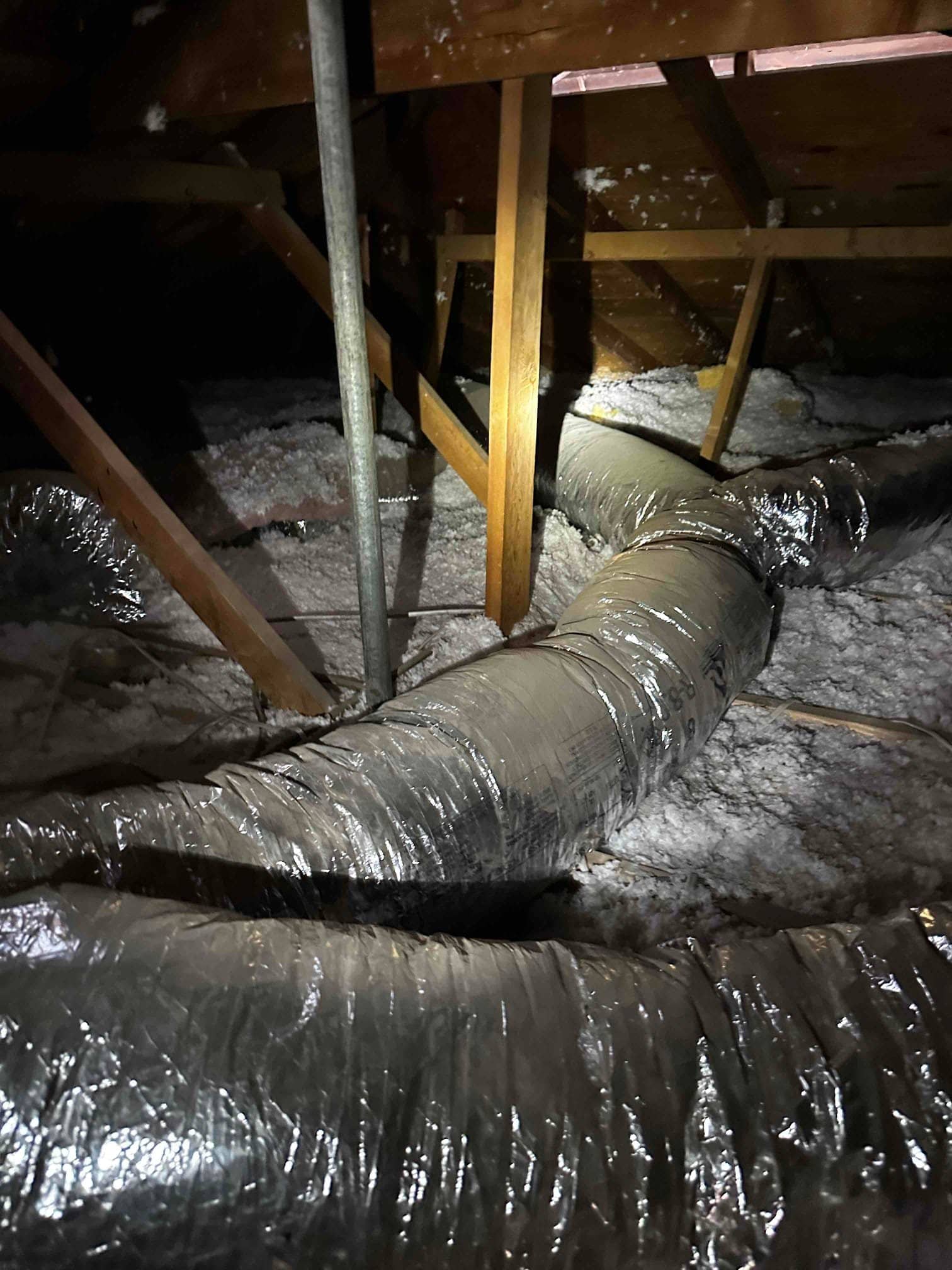 Flexible insulated ducts run through an attic with blown-in insulation, shown during installation with framing and roof decking visible.
