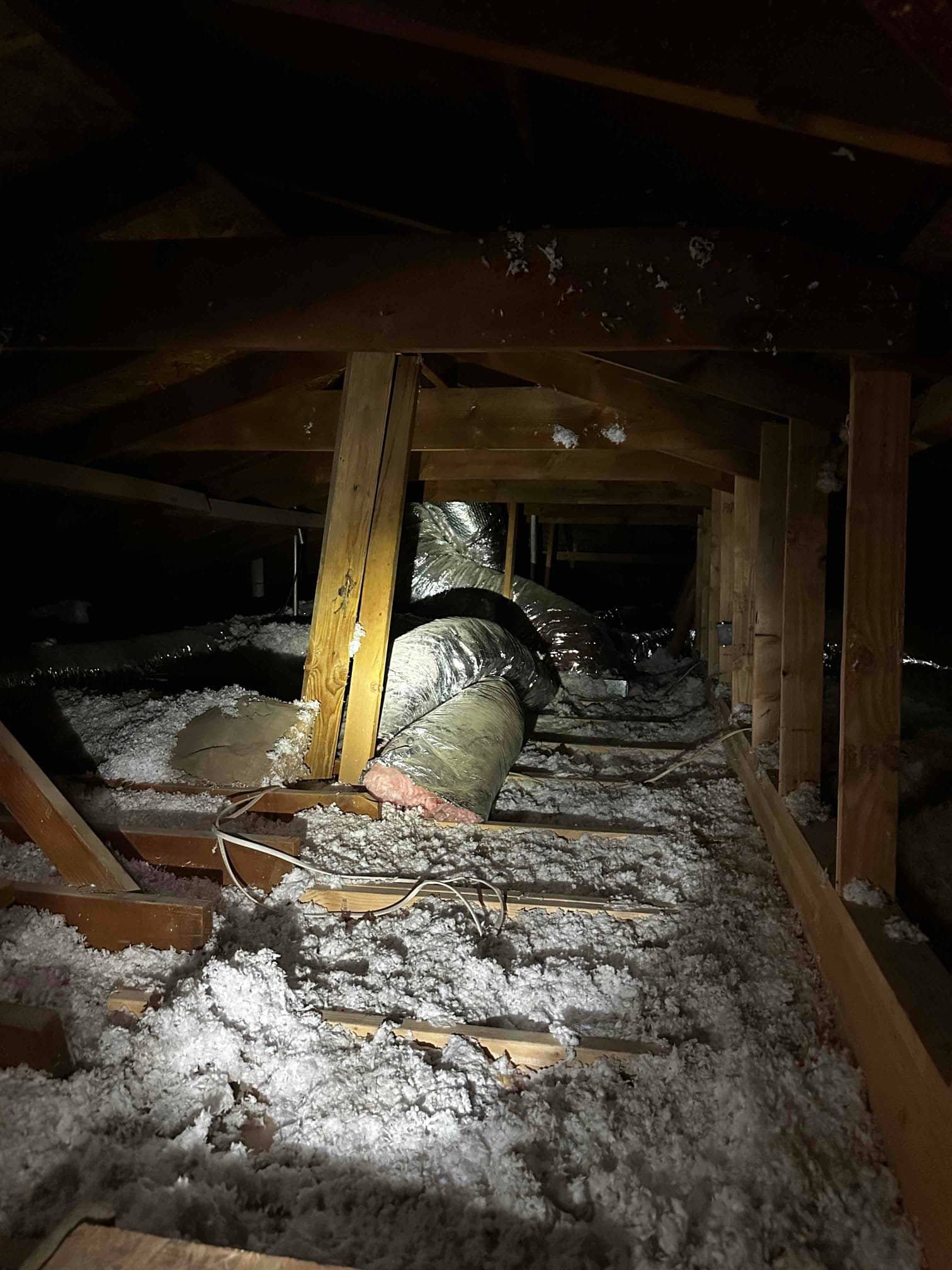 Flexible insulated ductwork routed through an attic with blown-in insulation and roof trusses illuminated by a work light.