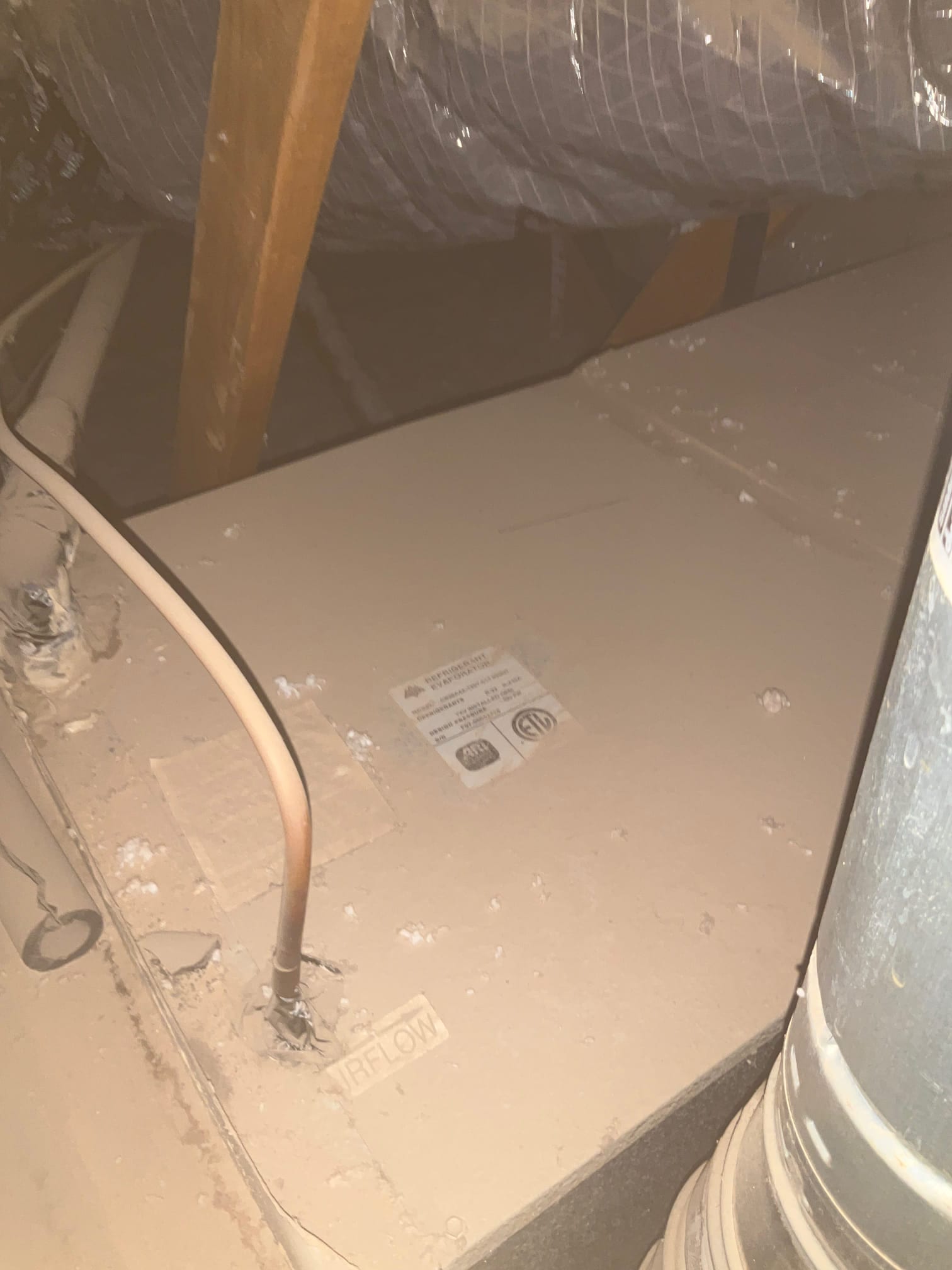 Attic air handler cabinet with insulated flex duct, PVC drain, and copper tubing; dusty top surface shows AIRFLOW marking.