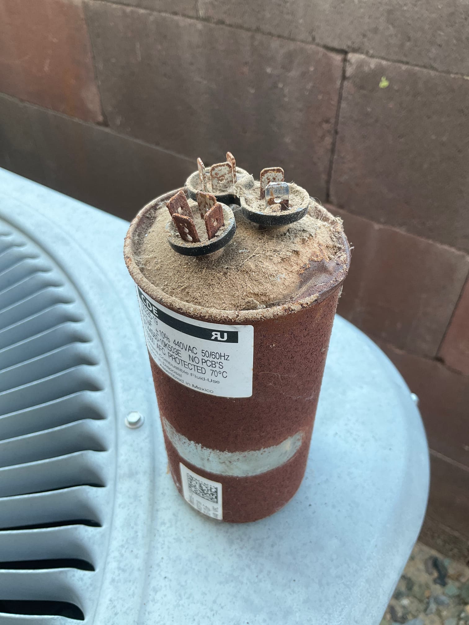 Dual-run capacitor removed and placed on condenser top, showing heavy rust, dust, and corroded terminals.