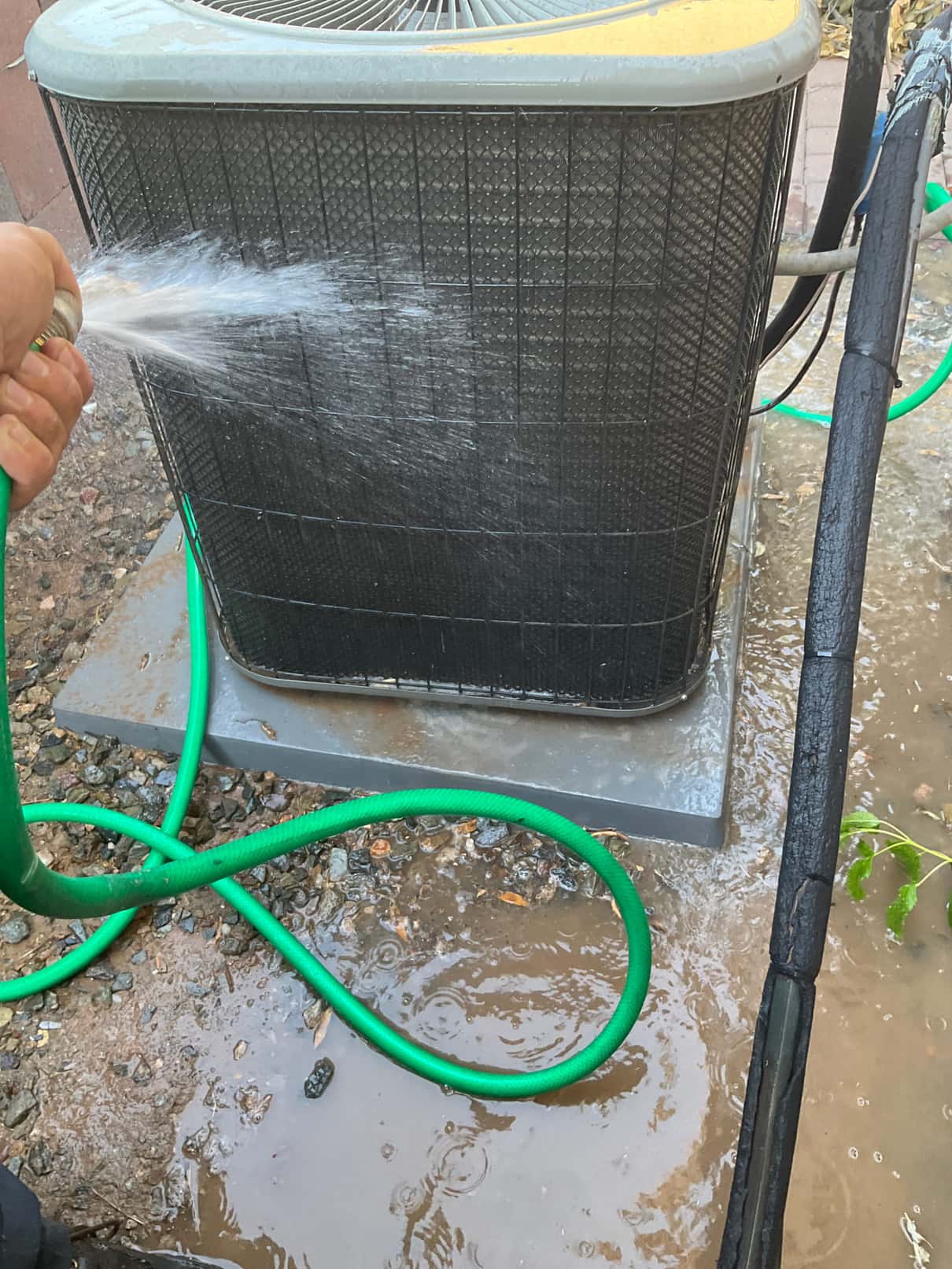 Outdoor condenser coil is rinsed with a garden hose; water pools around the base and pad.