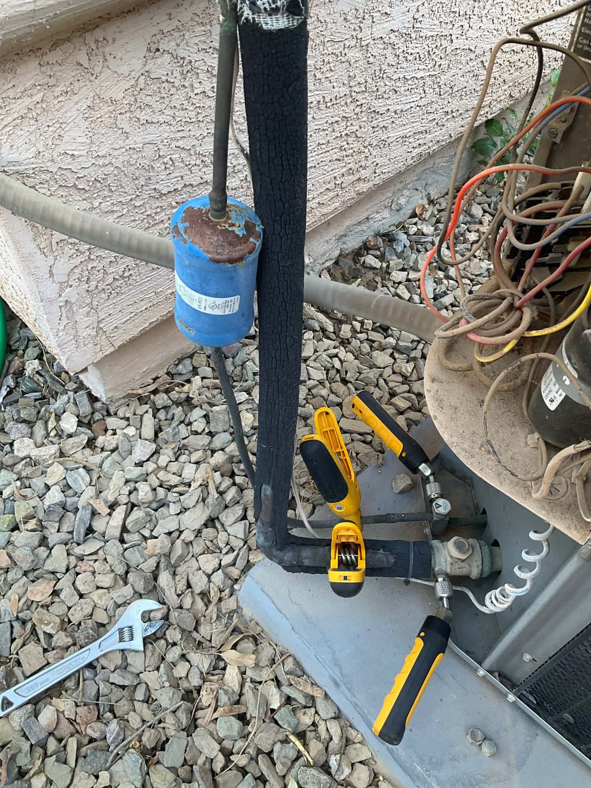 Outdoor condenser service area showing refrigerant lines with insulation, a rusted blue filter-drier, and two yellow clamps positioned on the lines.