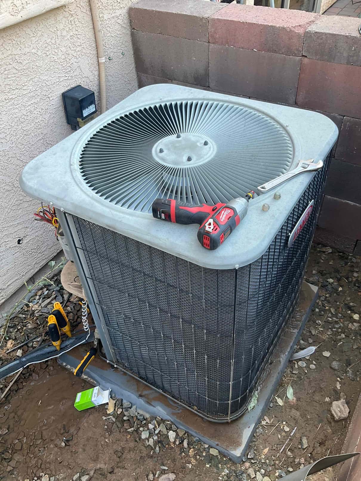 Outdoor condenser unit with fan grille on top, side panel open with wiring exposed, tools and clamps present.
