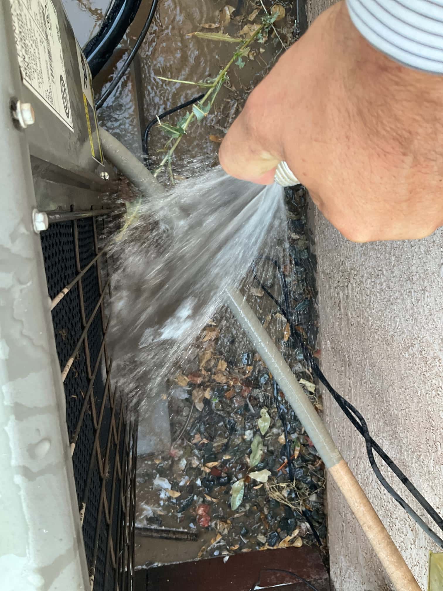 Water from a hose sprays the side condenser coil area, with insulated refrigerant line and wet debris below.
