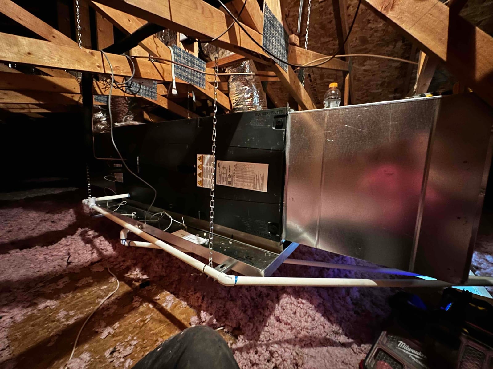 Air handler hung in attic with metal supply plenum, insulated ducts, condensate drain piping, and suspended support chains visible.