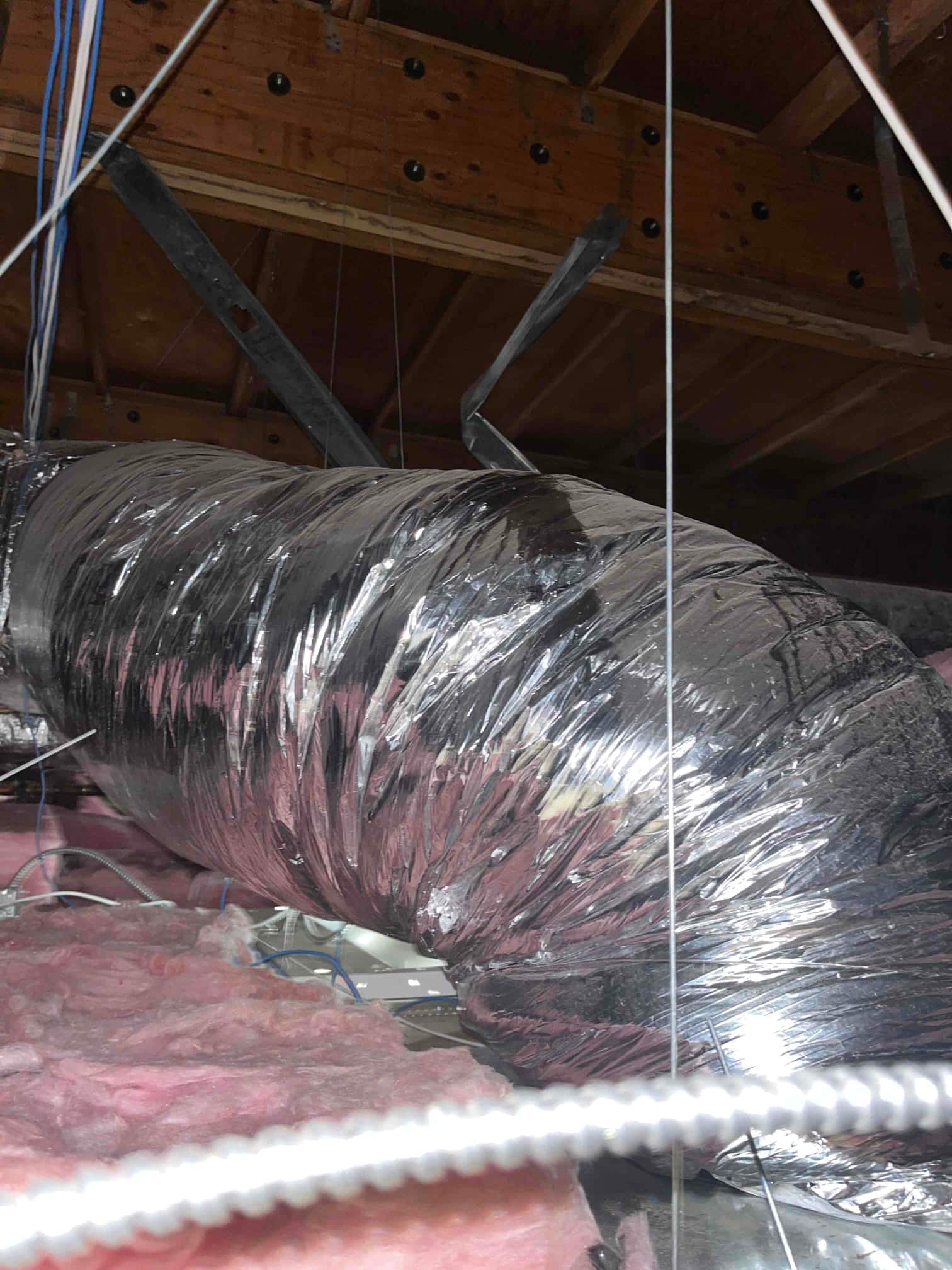 Flex duct in an attic with reflective insulation jacket, supported by straps, above exposed pink fiberglass batt insulation.