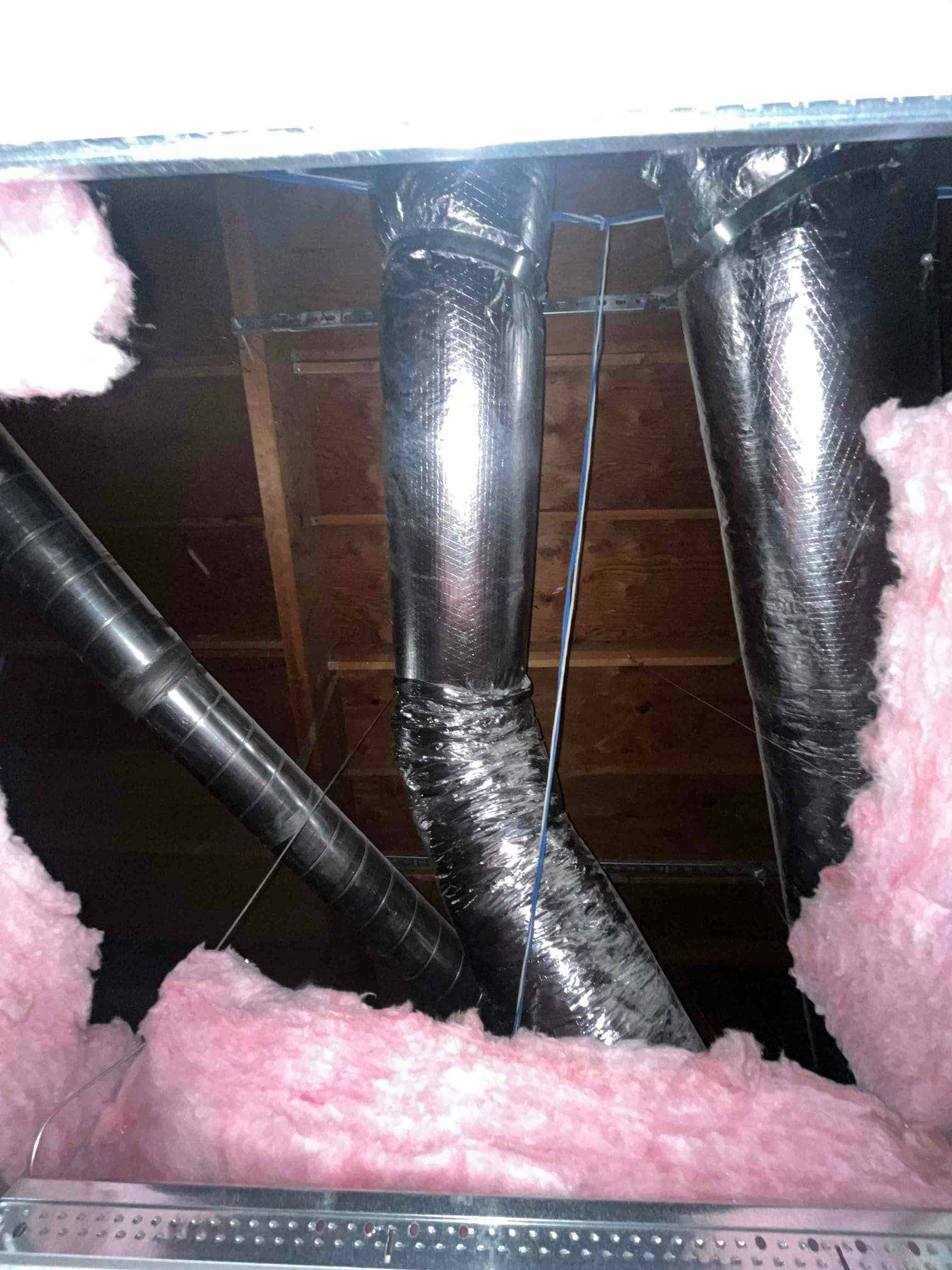 Flexible insulated ductwork and metal round ducting run through an attic space with exposed insulation and wooden roof decking.