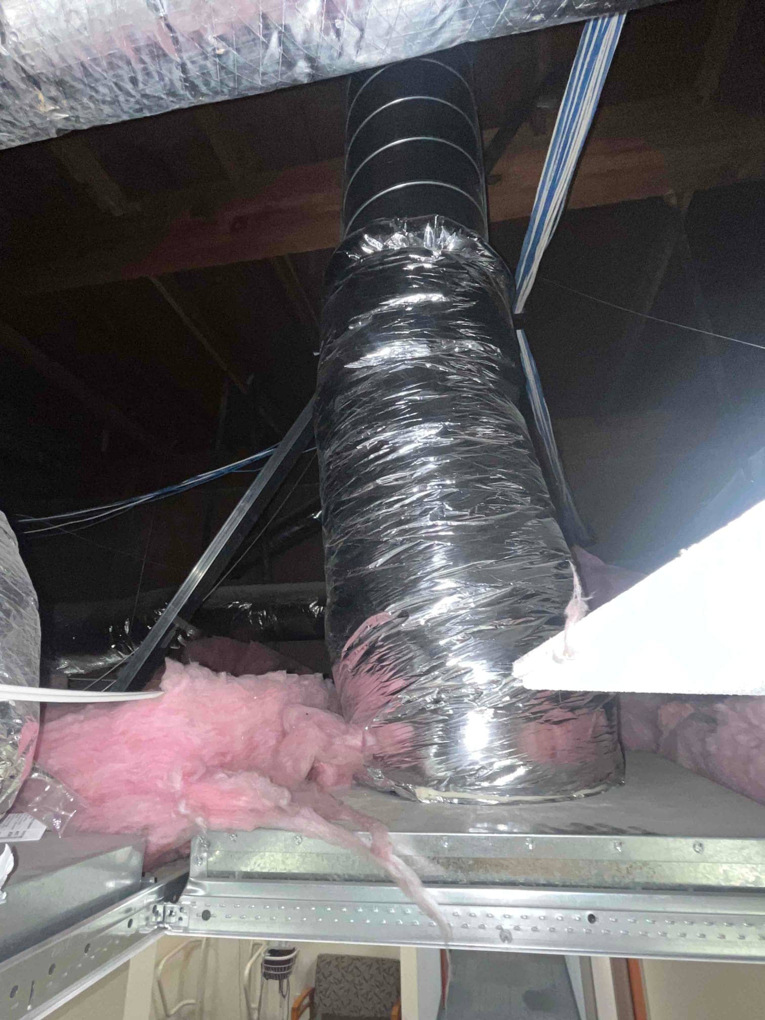 Flexible insulated ductwork runs through a ceiling space with exposed insulation and support strapping visible around the duct.
